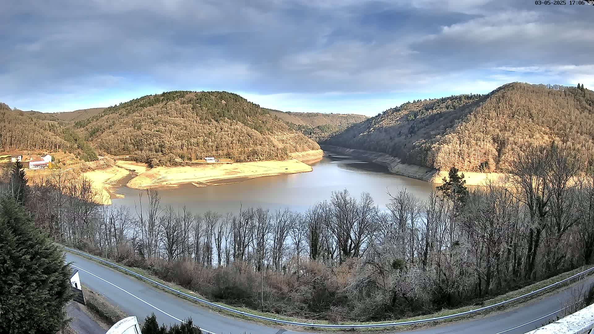 A partly cloudy day reveals a winding river nestled between hillsides covered in leafless trees, with a road curving around the lower right.