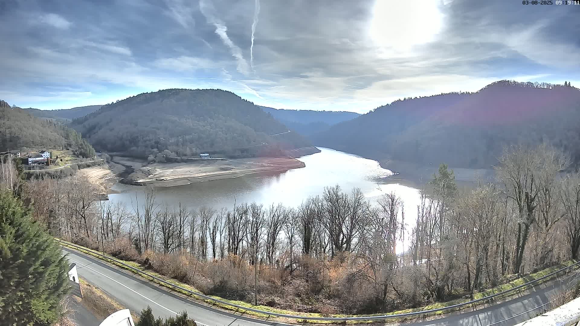 A partly cloudy day reveals a winding river valley with barren trees lining the shores and hillsides, a road curving into view, and a low water level in the reservoir.