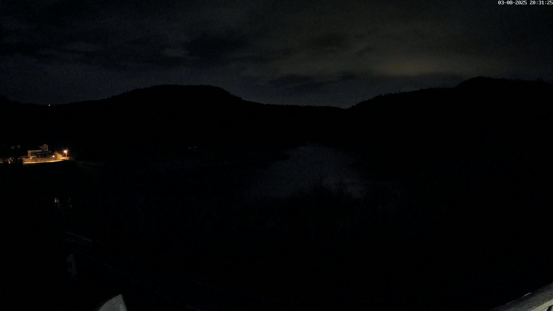 A nighttime view of dark, silhouetted hills and a body of water under a cloudy sky, with a single illuminated building visible in the distance.