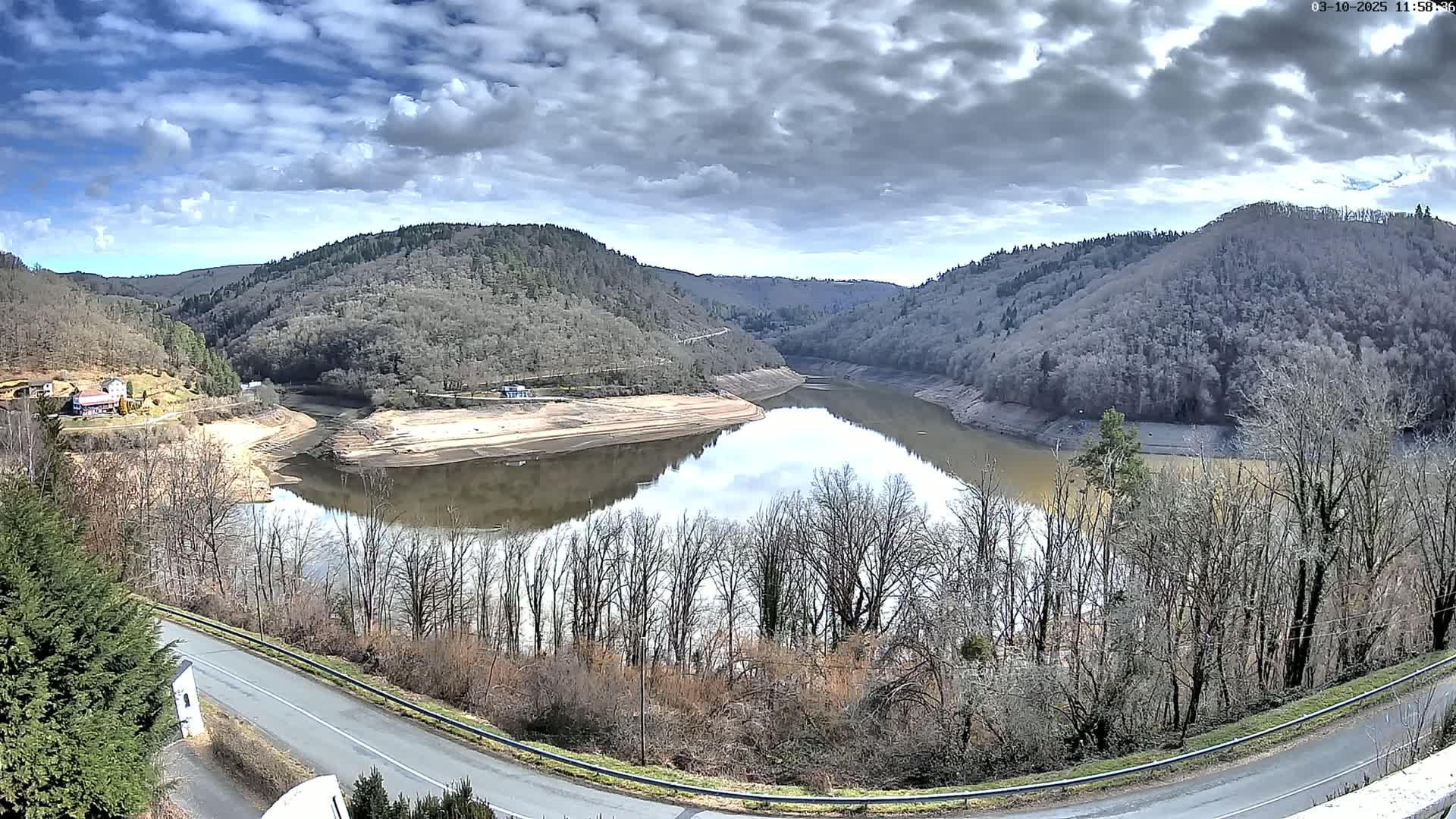 A partly cloudy day reveals a winding reservoir nestled in a valley between barren, tree-covered hills, with a road curving along the lower edge of the scene.