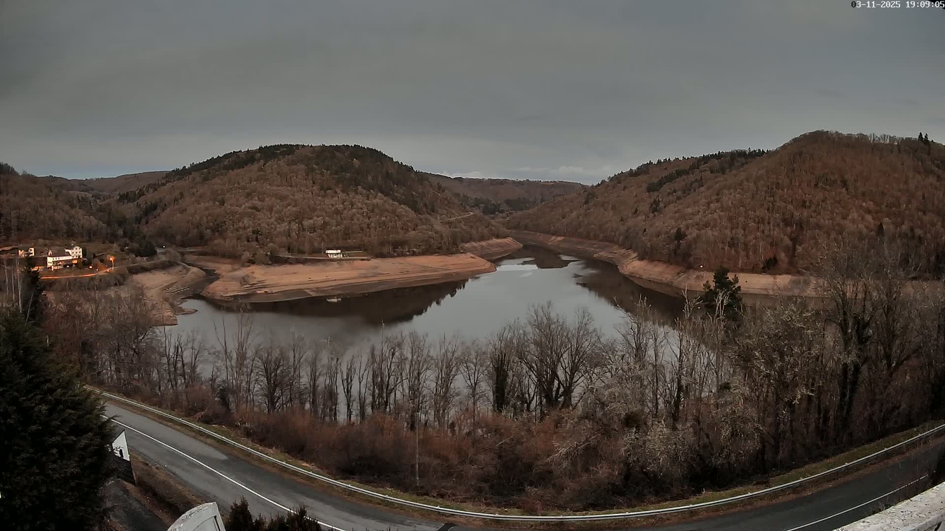 A calm, dark-colored lake nestled in a valley between hills of bare, brown trees under a cloudy sky.