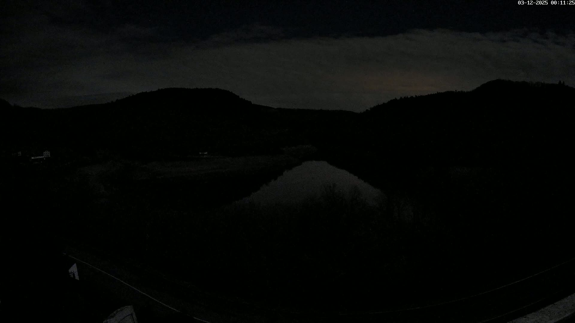 A nighttime view of a dark, still body of water nestled between silhouetted hills under a cloudy sky.
