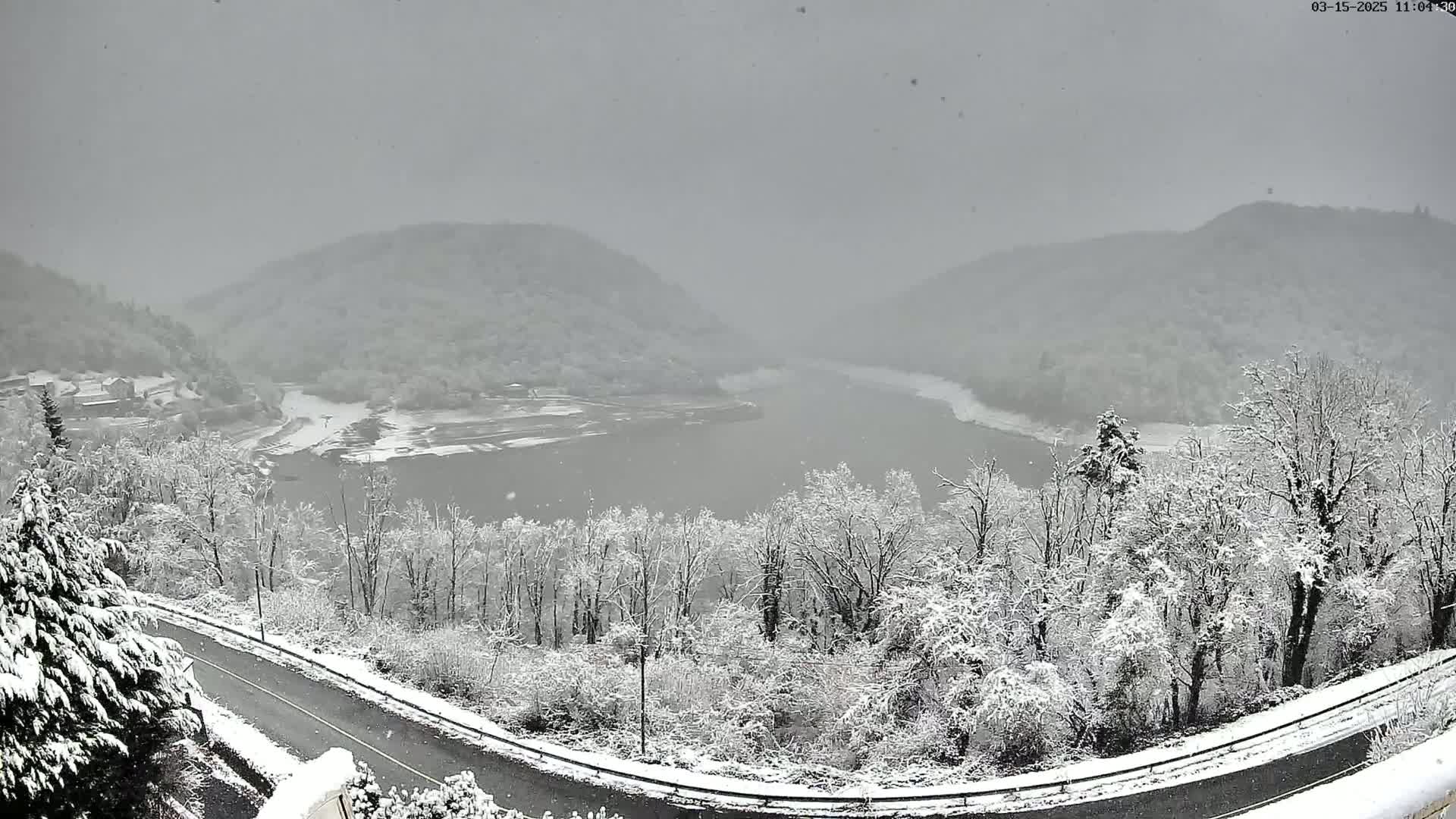 A snow-covered landscape shows a winding road overlooking a lake nestled between snow-dusted hills, with light snowfall continuing.
