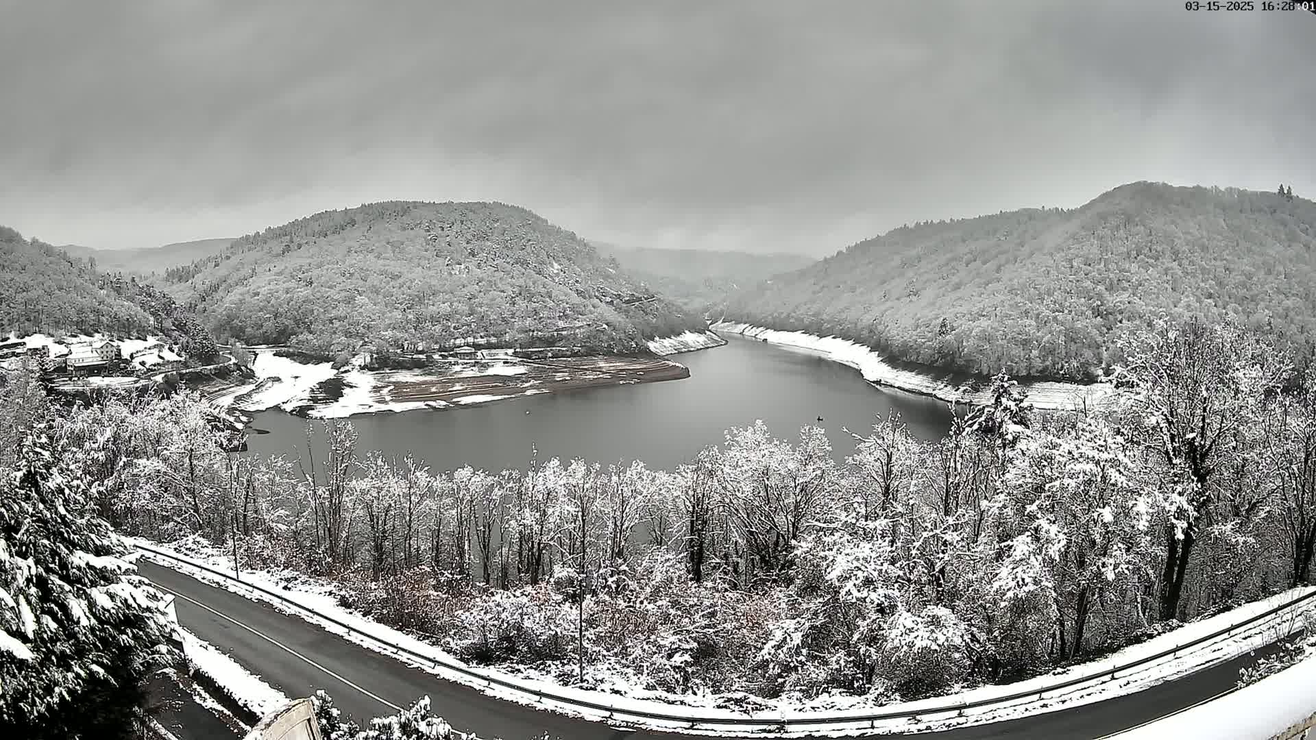 A snow-covered landscape featuring a winding road, a lake nestled between snow-dusted hills, and a overcast sky.