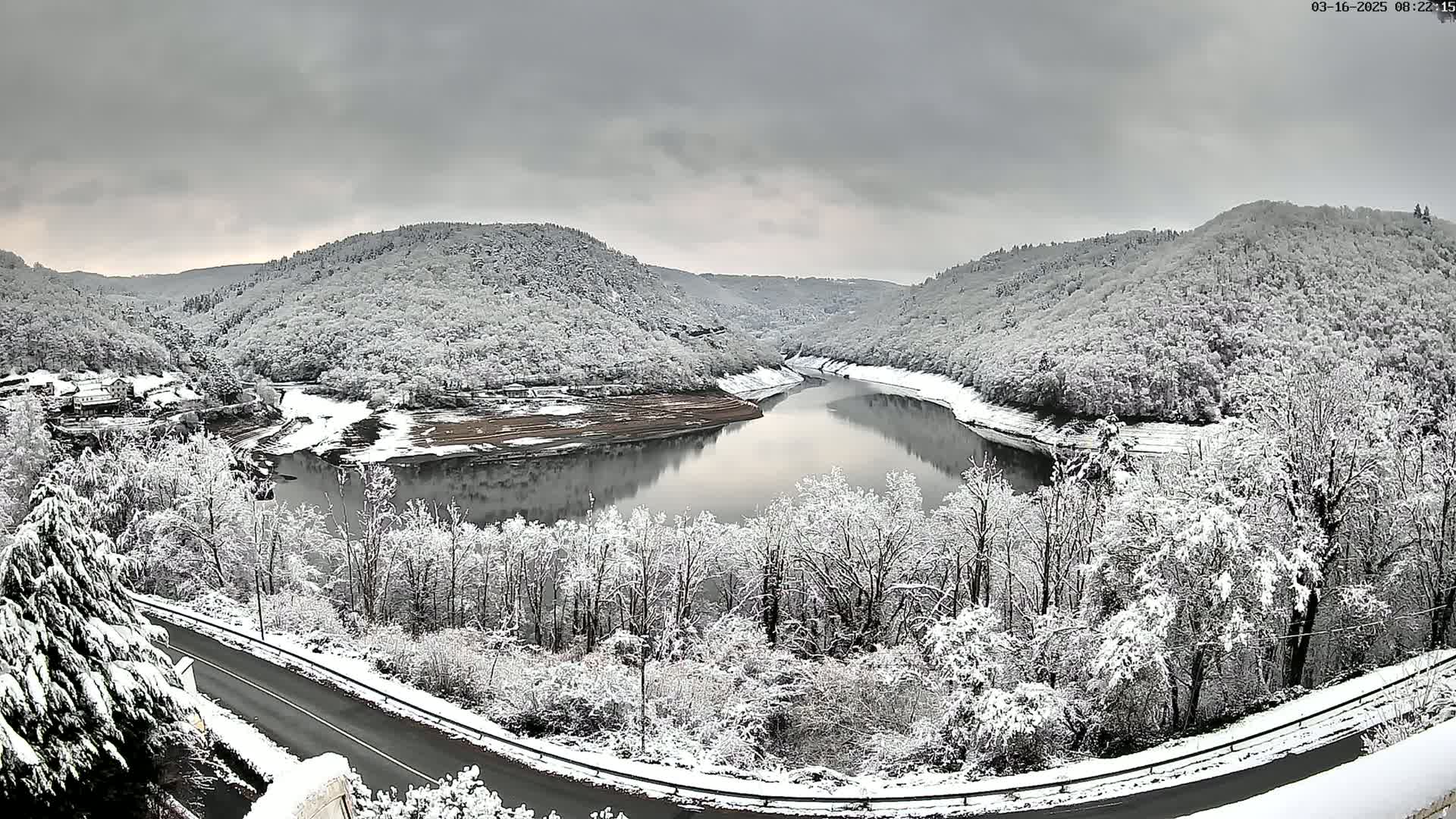 A snow-covered landscape featuring a winding river reflecting the overcast sky, surrounded by snow-laden hills and trees, with a road curving in the foreground.