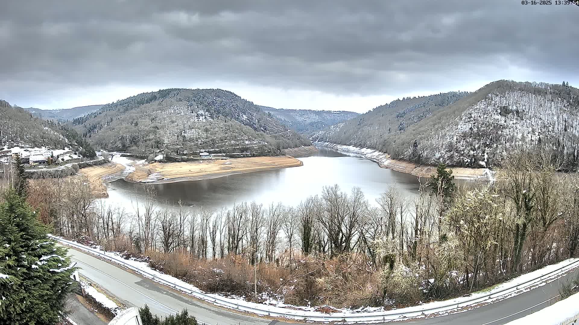 A reservoir curves through a valley under a cloudy sky, with snow dusting the hillsides and ground.
