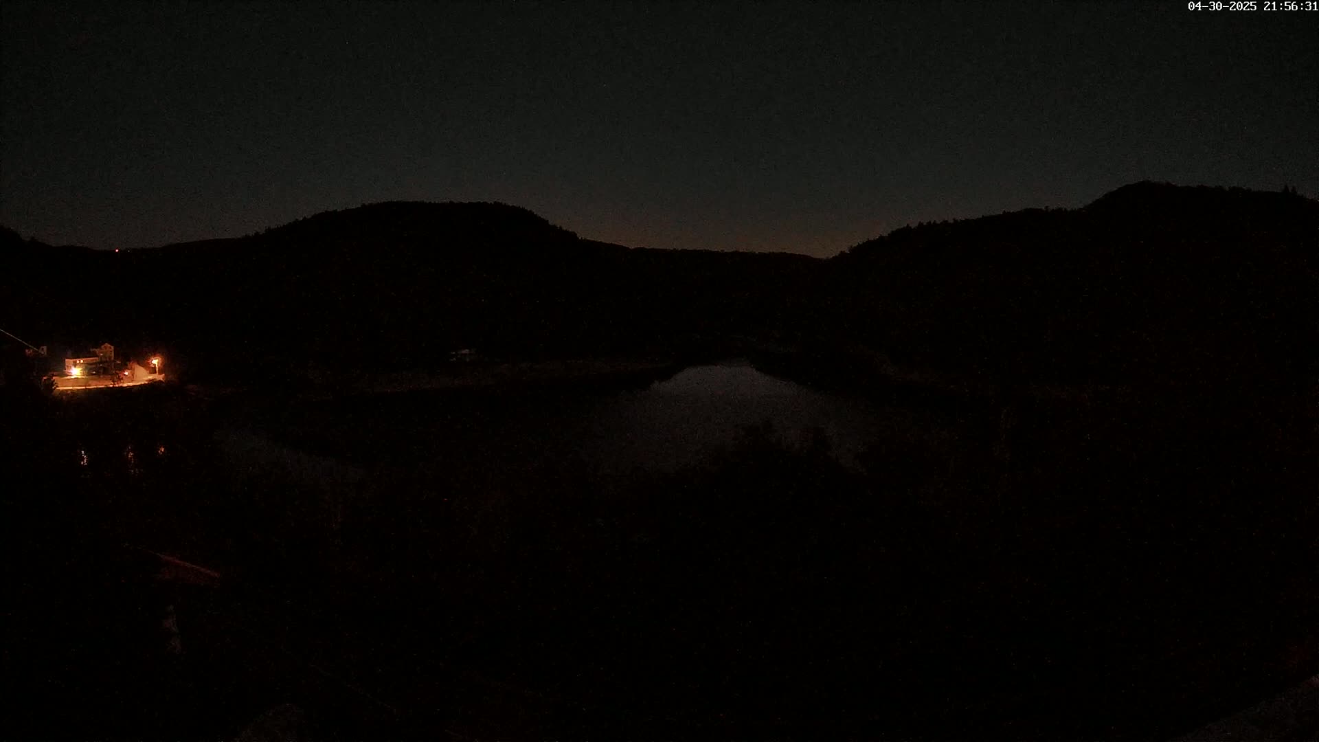 A dark, nighttime view shows a body of water nestled between hills, with a single light visible near the water's edge.