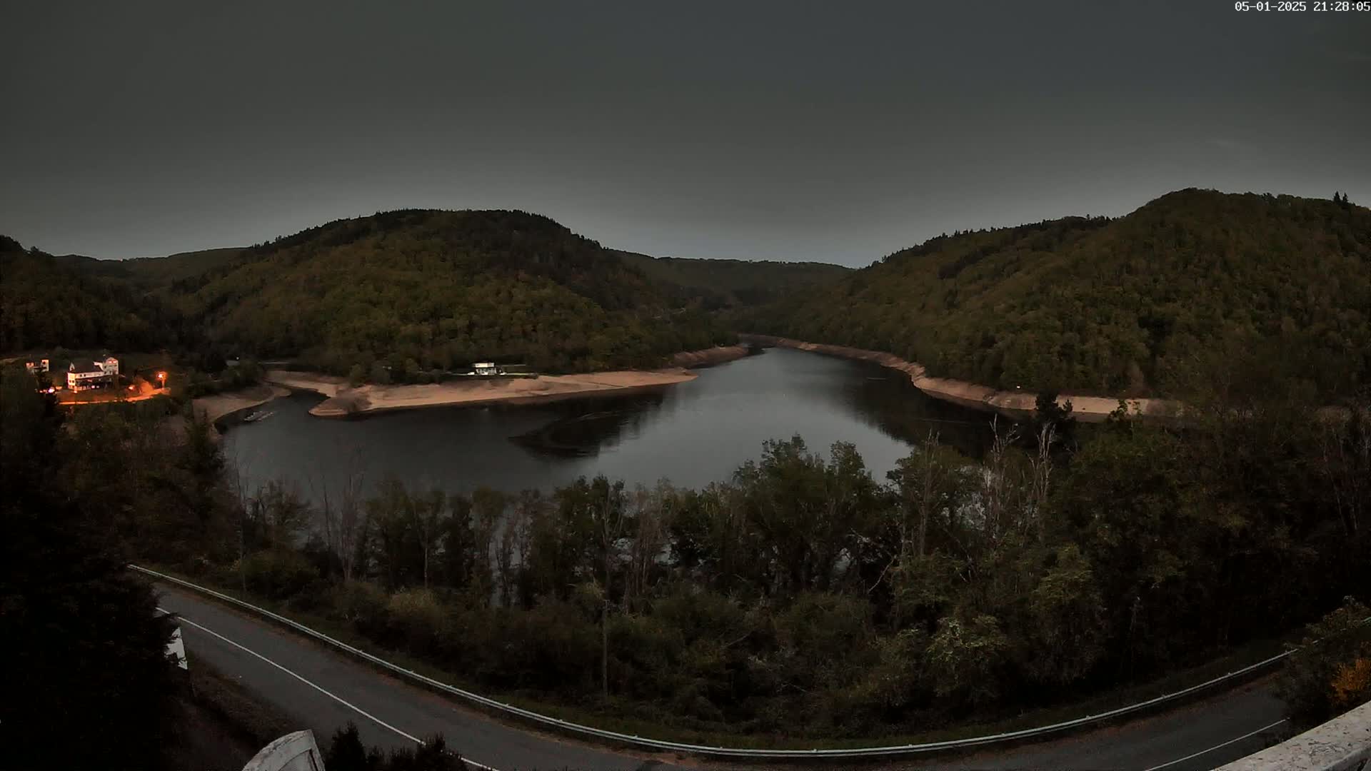 A dark, winding river flows between tree-covered hills under a dusky sky.