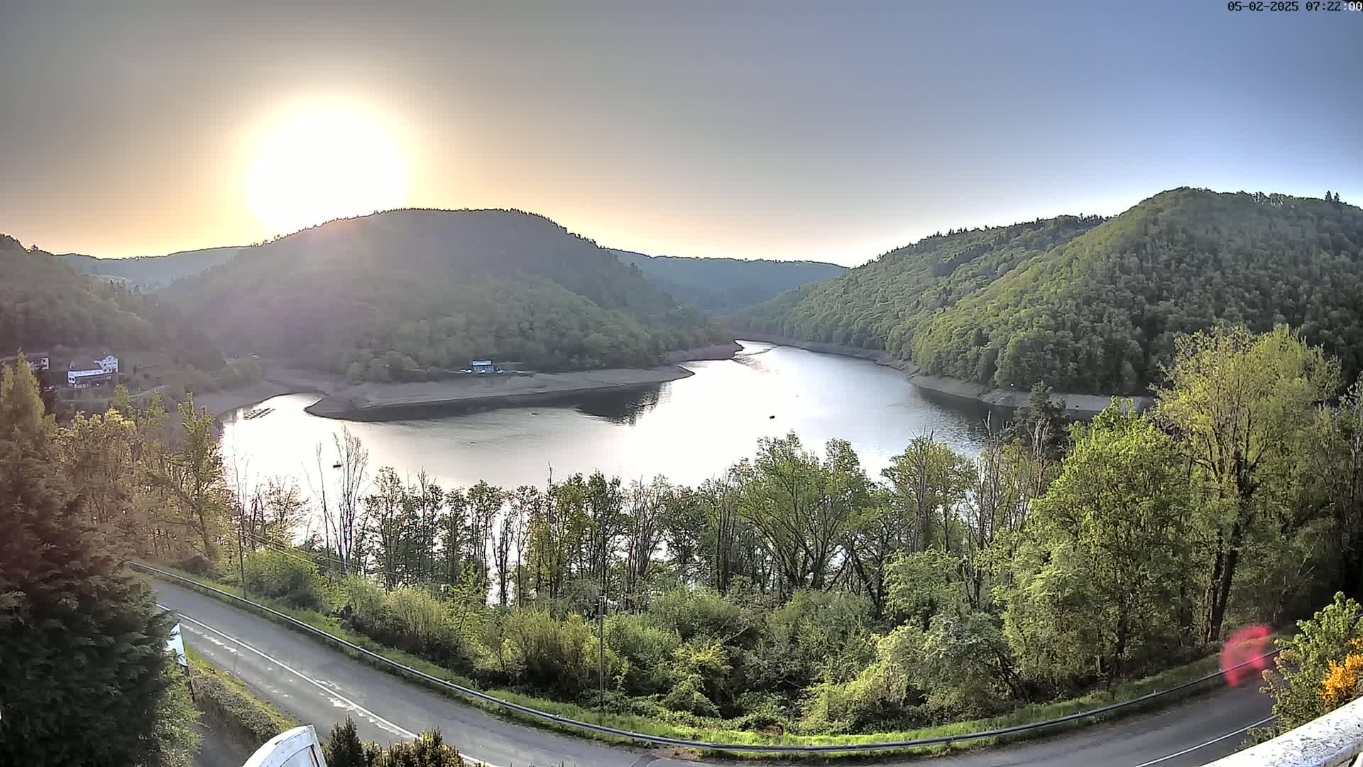 A calm lake nestled between verdant hills under a bright, setting sun.