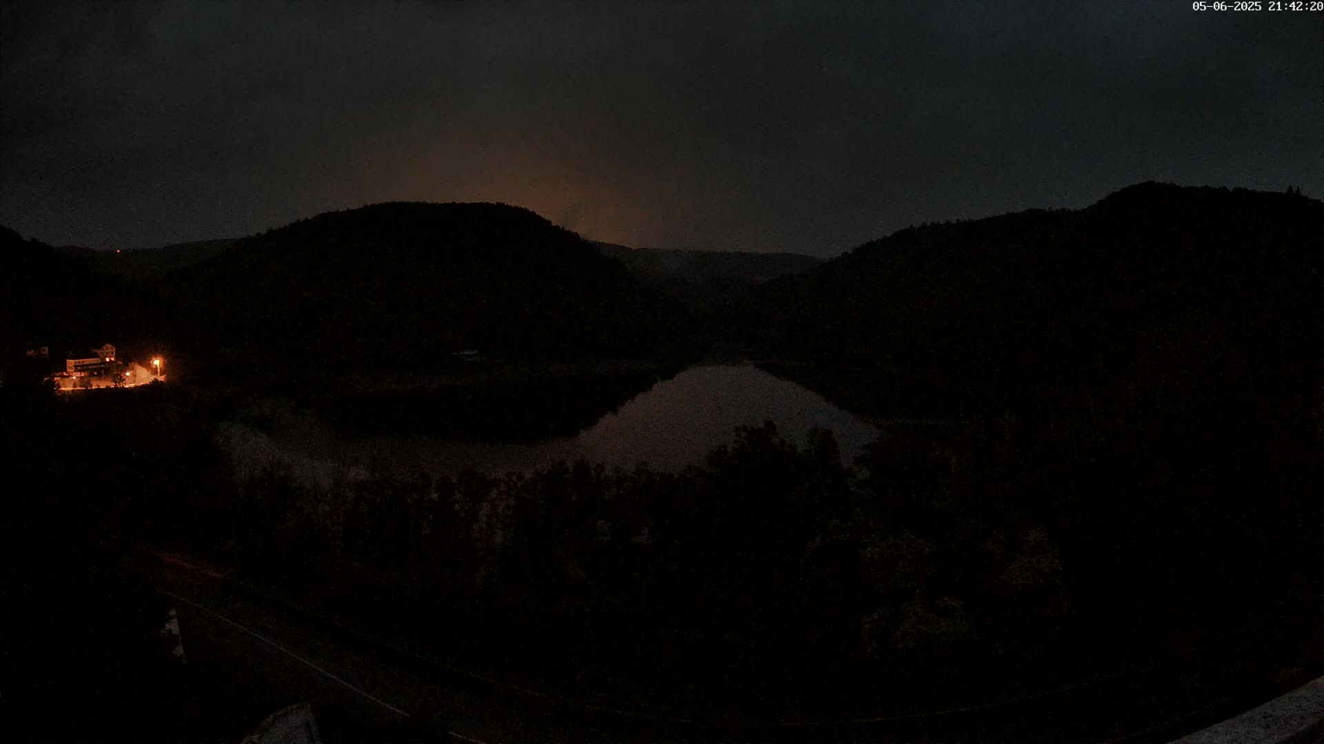 A dark, nighttime scene shows a calm body of water nestled between two hills, with a single illuminated building visible on the left.