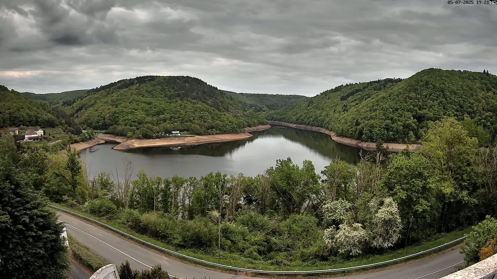 A curved lake nestled in a valley of lush green hills under a cloudy sky.