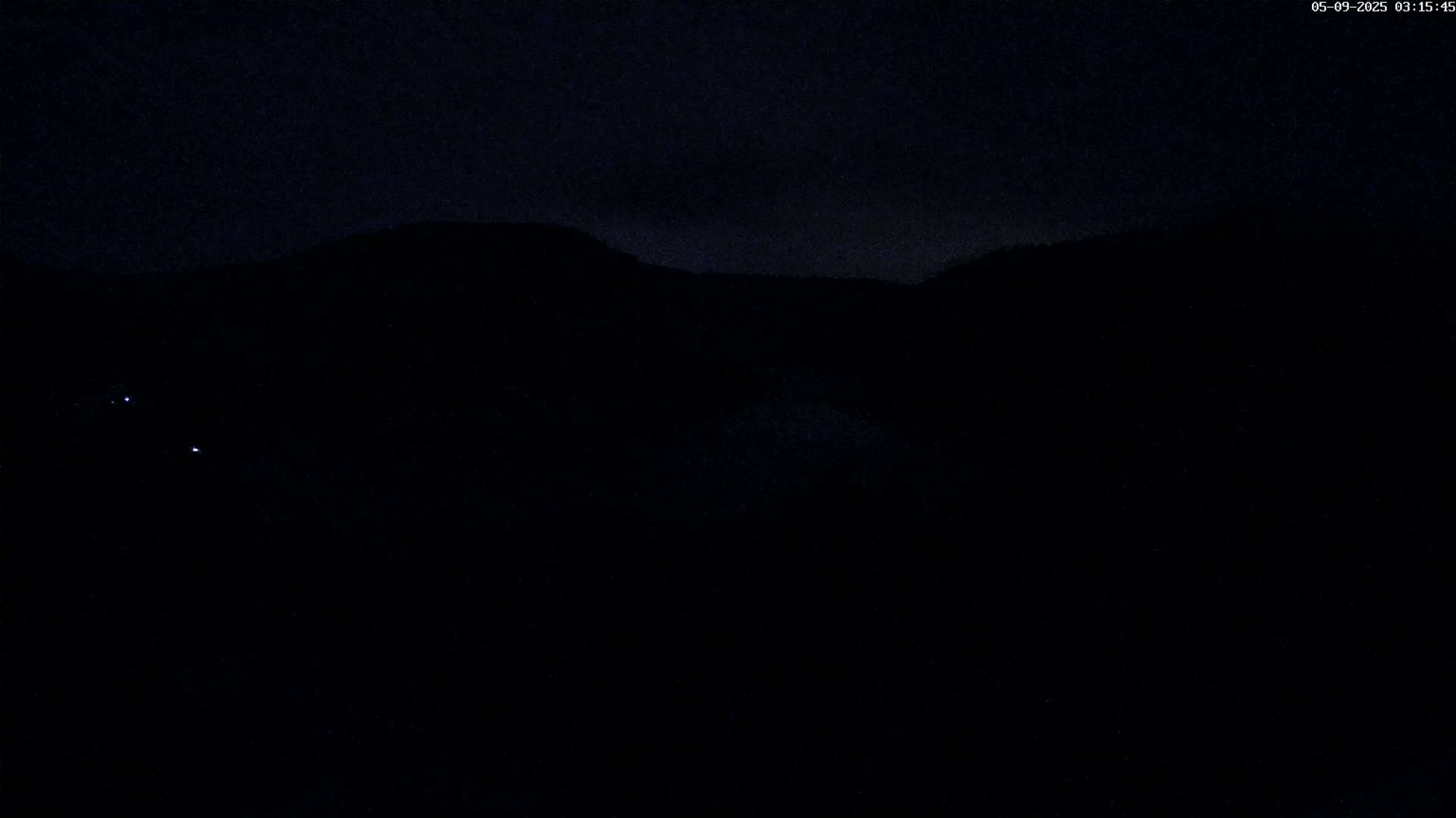 A nighttime view of a dark, silhouetted landscape with a few faint lights visible in the distance under a cloudy sky.