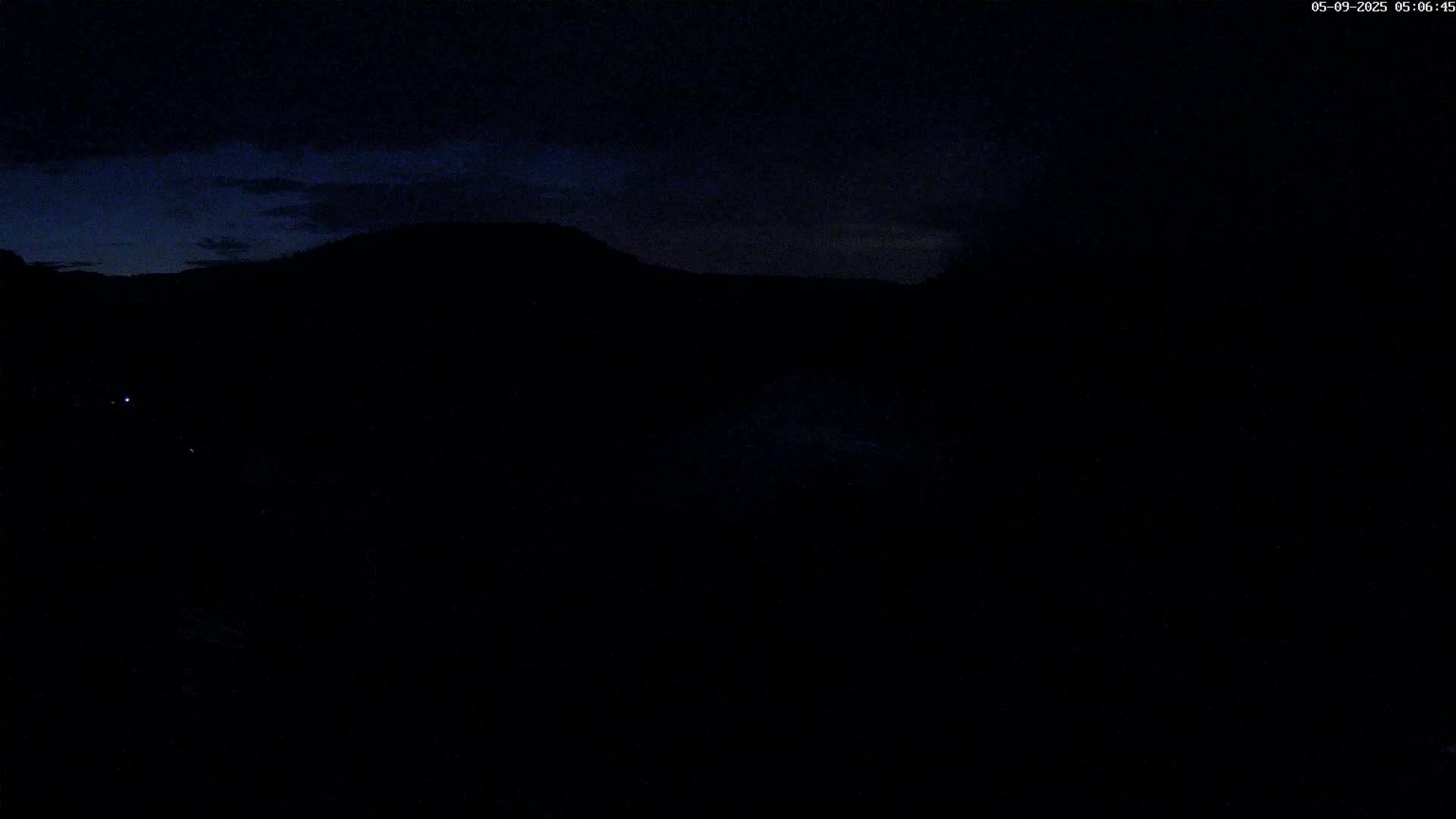 A dark, nighttime landscape shows a silhouetted mountain range under a mostly clear, dark blue sky.