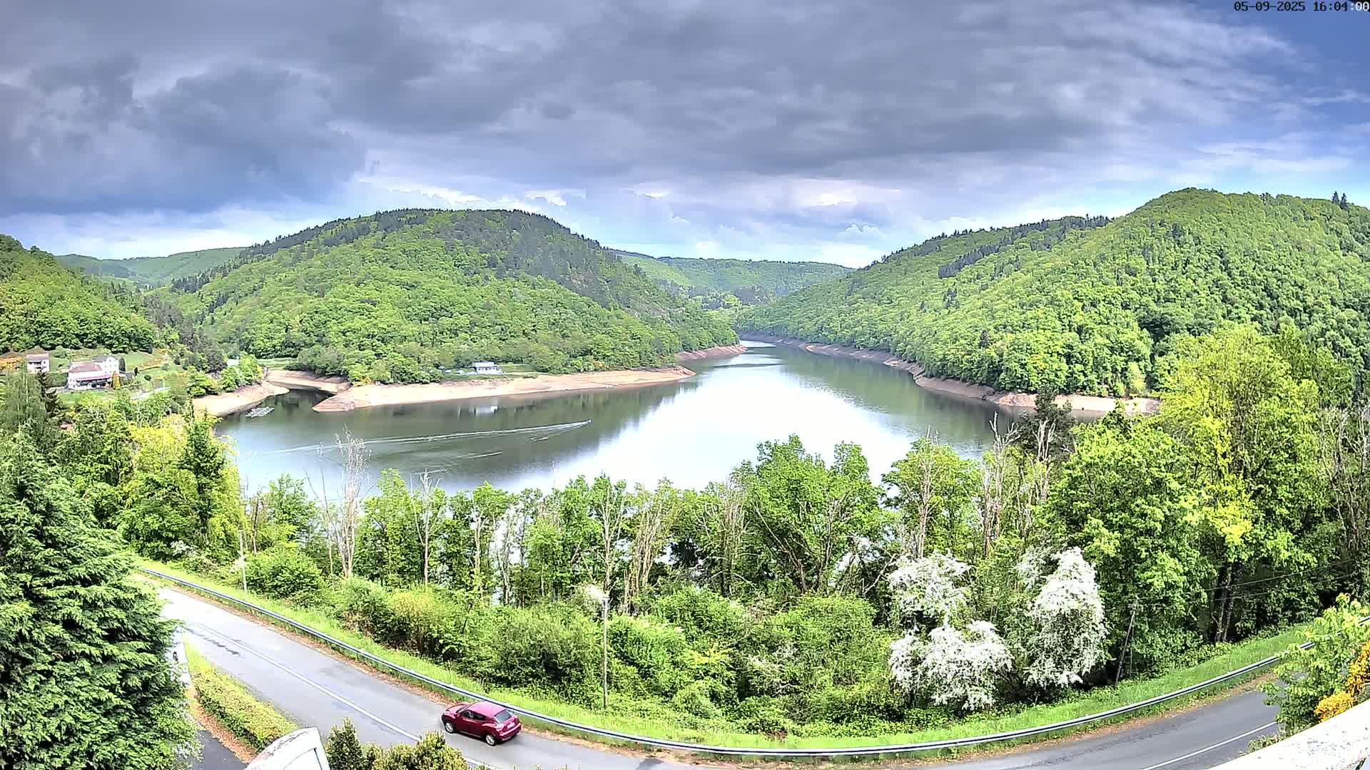 A red car drives on a road overlooking a calm lake nestled in a valley of lush green hills under a cloudy sky.