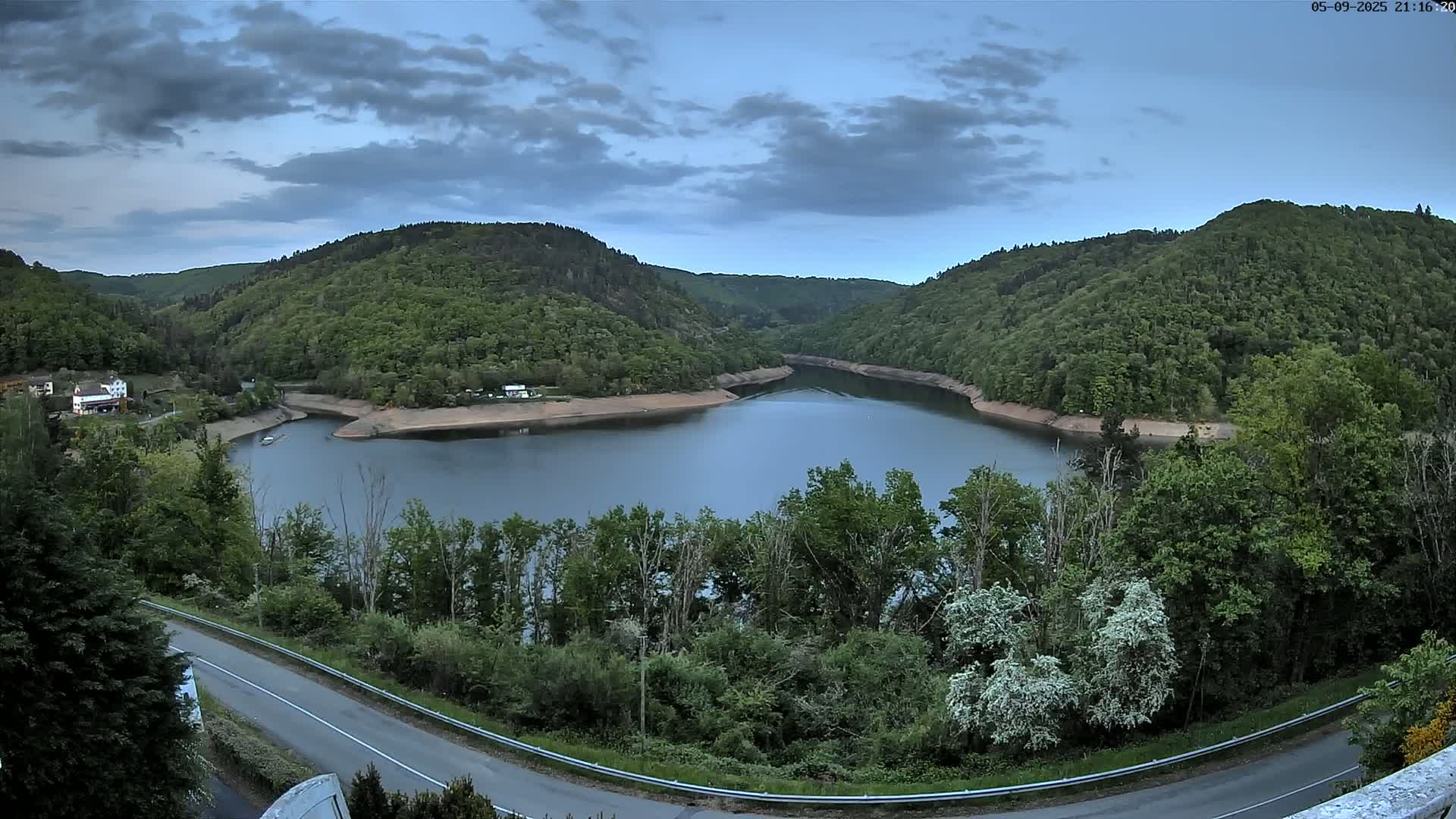 A crescent-shaped lake nestled in a valley of lush green hills under a partly cloudy sky.