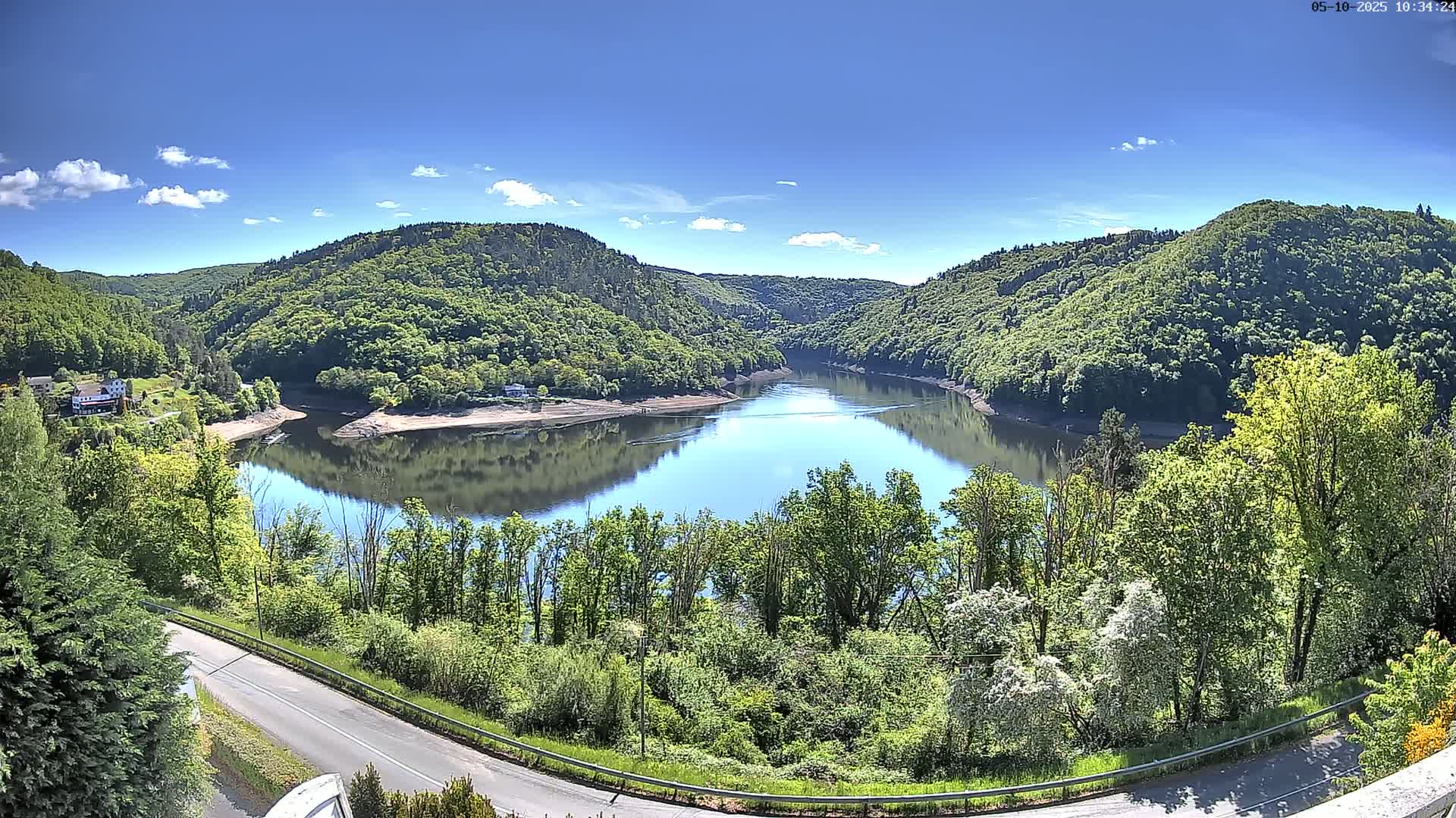 A calm lake nestled in a valley of lush green hills under a mostly sunny sky.