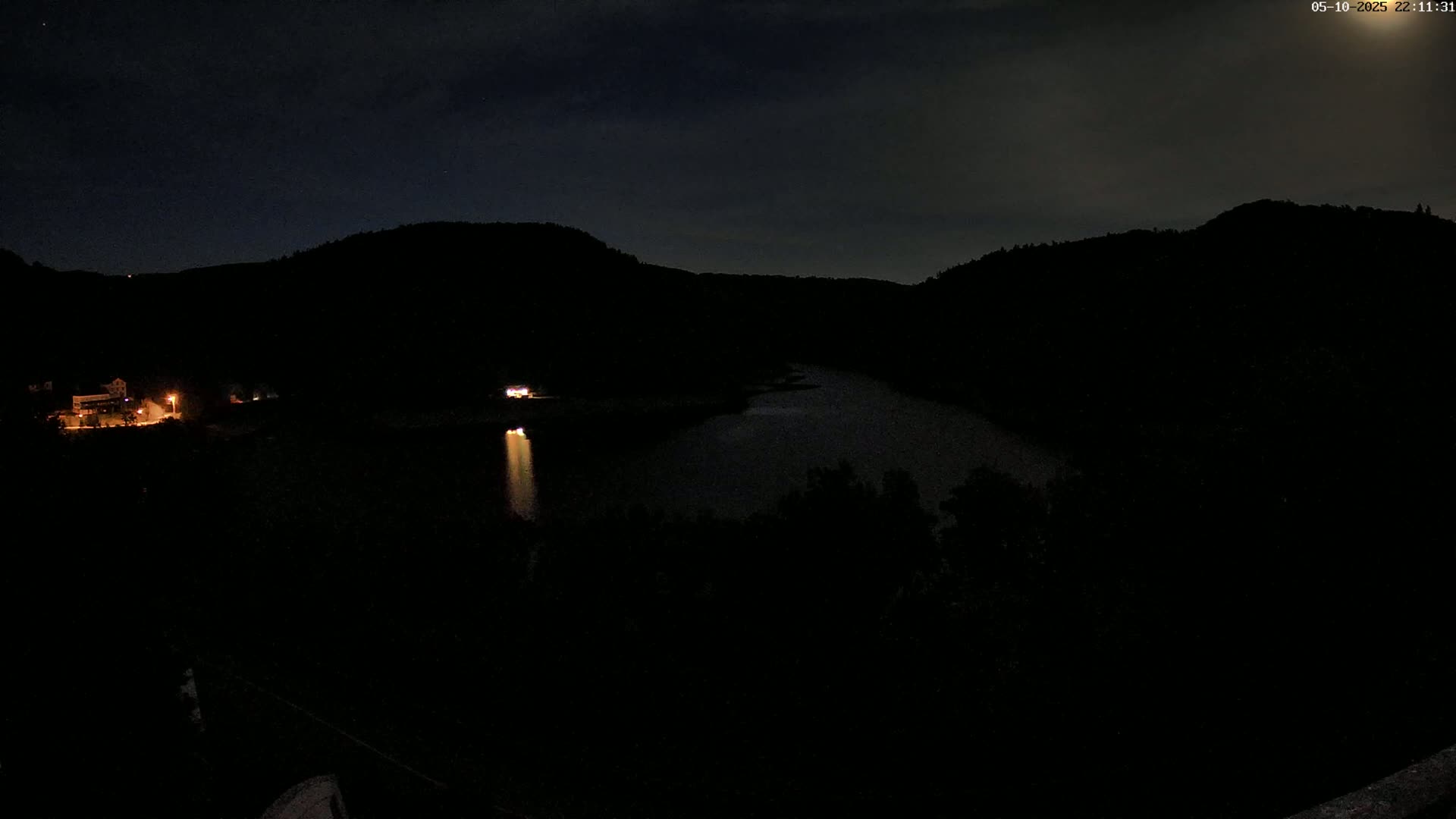A dark, nighttime view shows a calm body of water between silhouetted hills under a mostly cloudy sky.