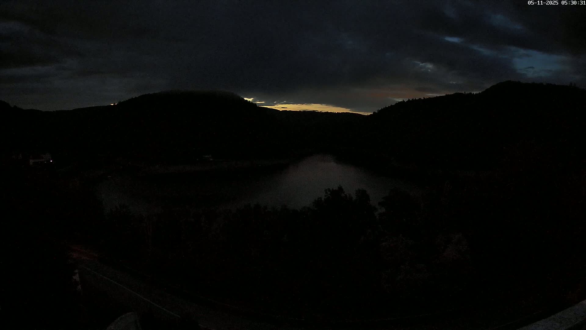 A dark, cloudy twilight scene shows a body of water nestled between silhouetted hills, with a sliver of light visible in the sky above.