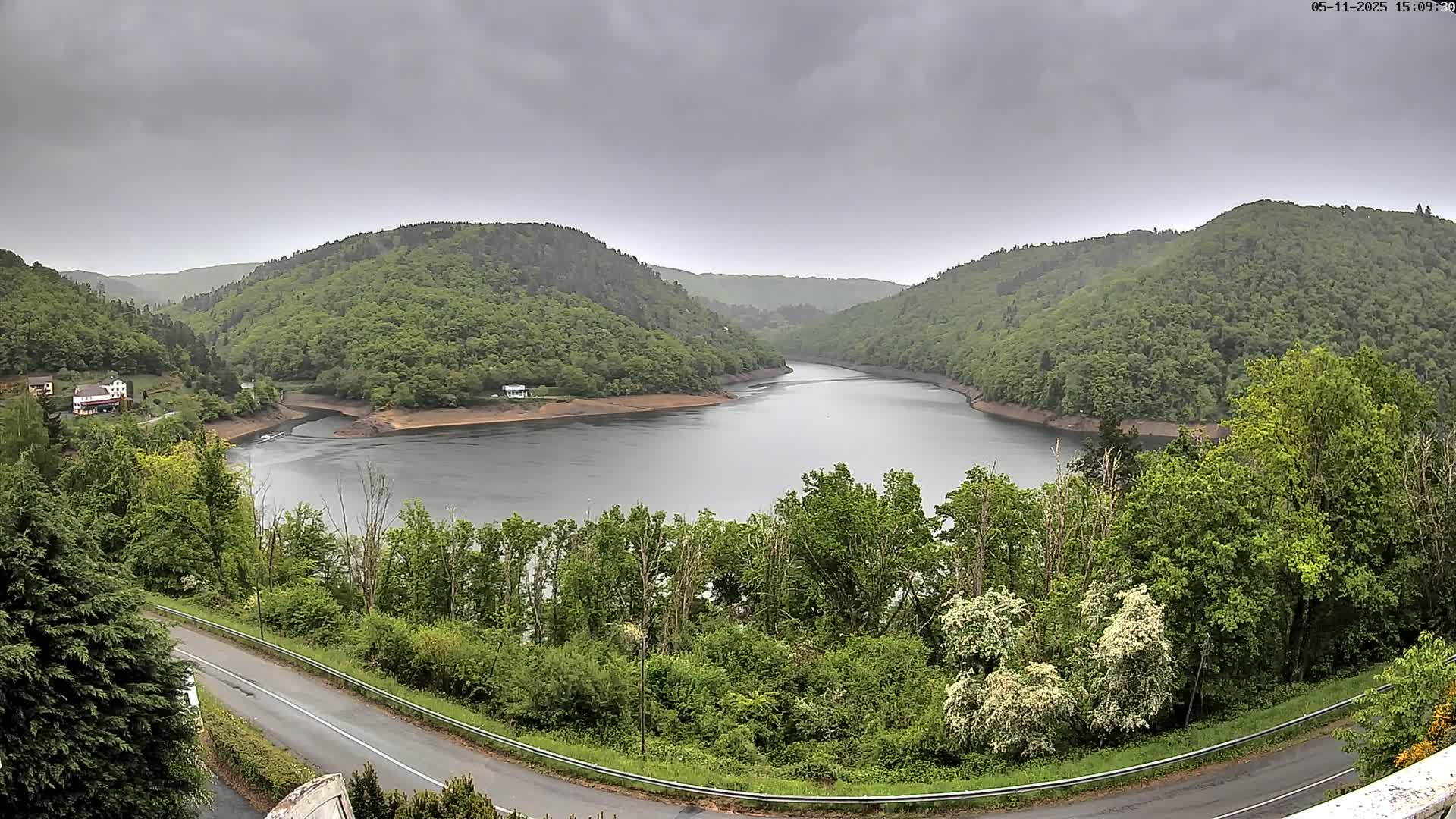 A winding river nestled in a valley of lush green hills under a cloudy sky is viewed from an elevated perspective, with a road curving along the valley floor.