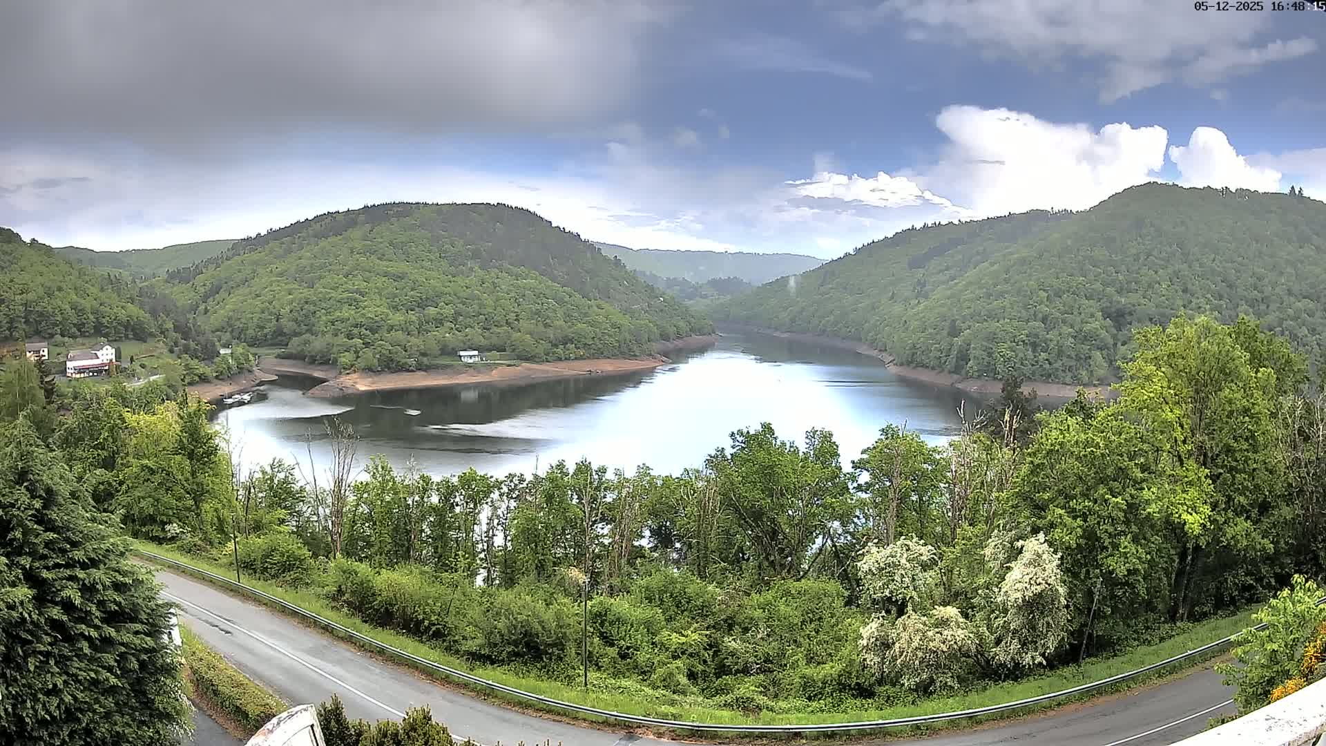 A partly cloudy sky overlooks a calm lake nestled between verdant hills, with a road curving alongside the lower hillside.
