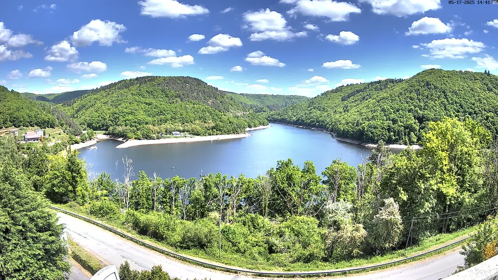 A curved lake nestled within a valley of lush green hills under a mostly sunny sky with scattered clouds.