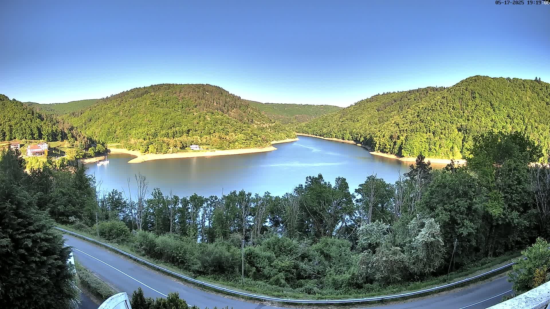 A tranquil lake nestled between lush green hills under a clear blue sky curves around a bend in the landscape near a road.