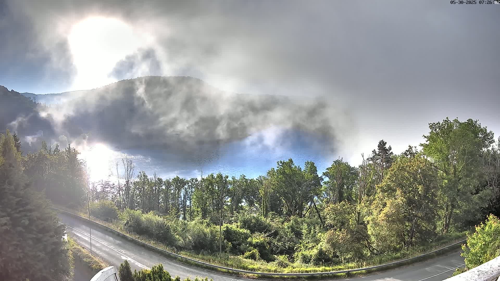 A partly sunny day with low-hanging fog obscures a lake nestled between tree-covered hills, with a winding road curving through the foreground.