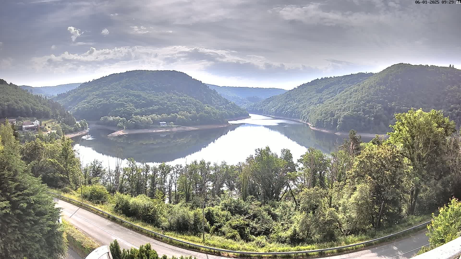 A calm lake nestled between lush green hills under a partly cloudy sky is viewed from a high vantage point above a winding road.
