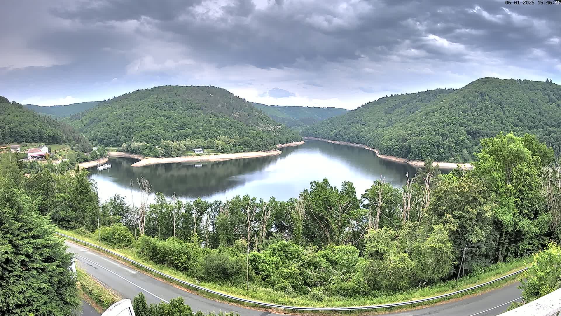 A winding road overlooks a calm lake nestled in a valley of lush green hills under a cloudy sky.