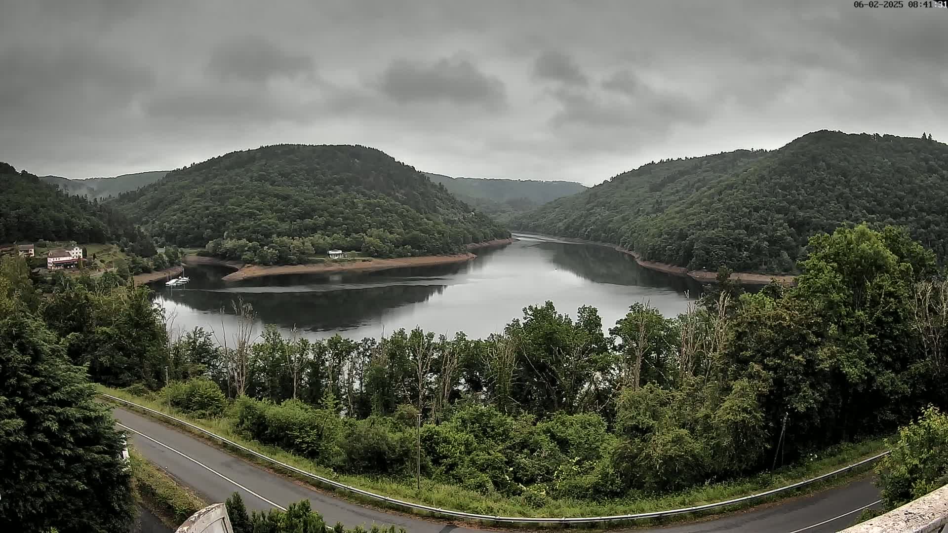 A calm lake nestled between lush green hills under a cloudy sky is seen from an elevated viewpoint, with a road curving below.