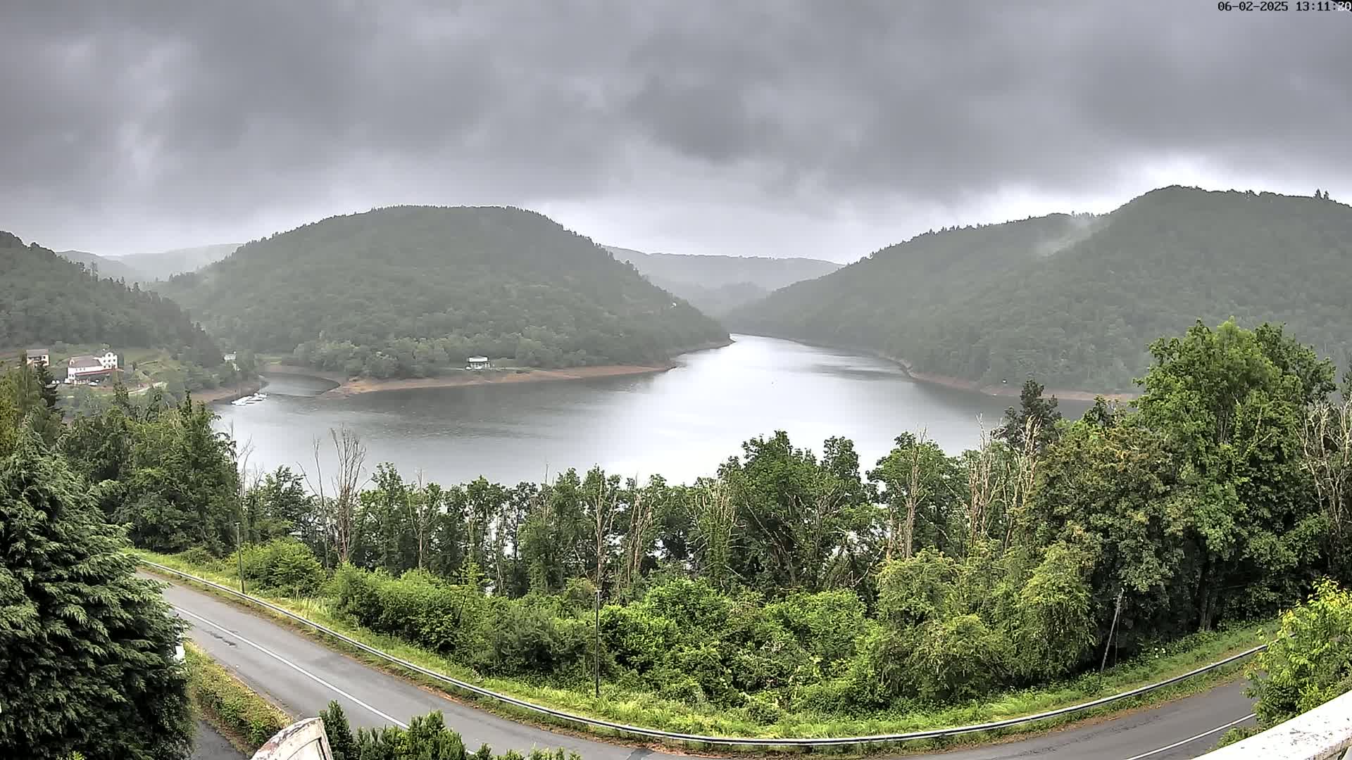 A winding road curves alongside a calm lake nestled between lush green hills under an overcast sky.