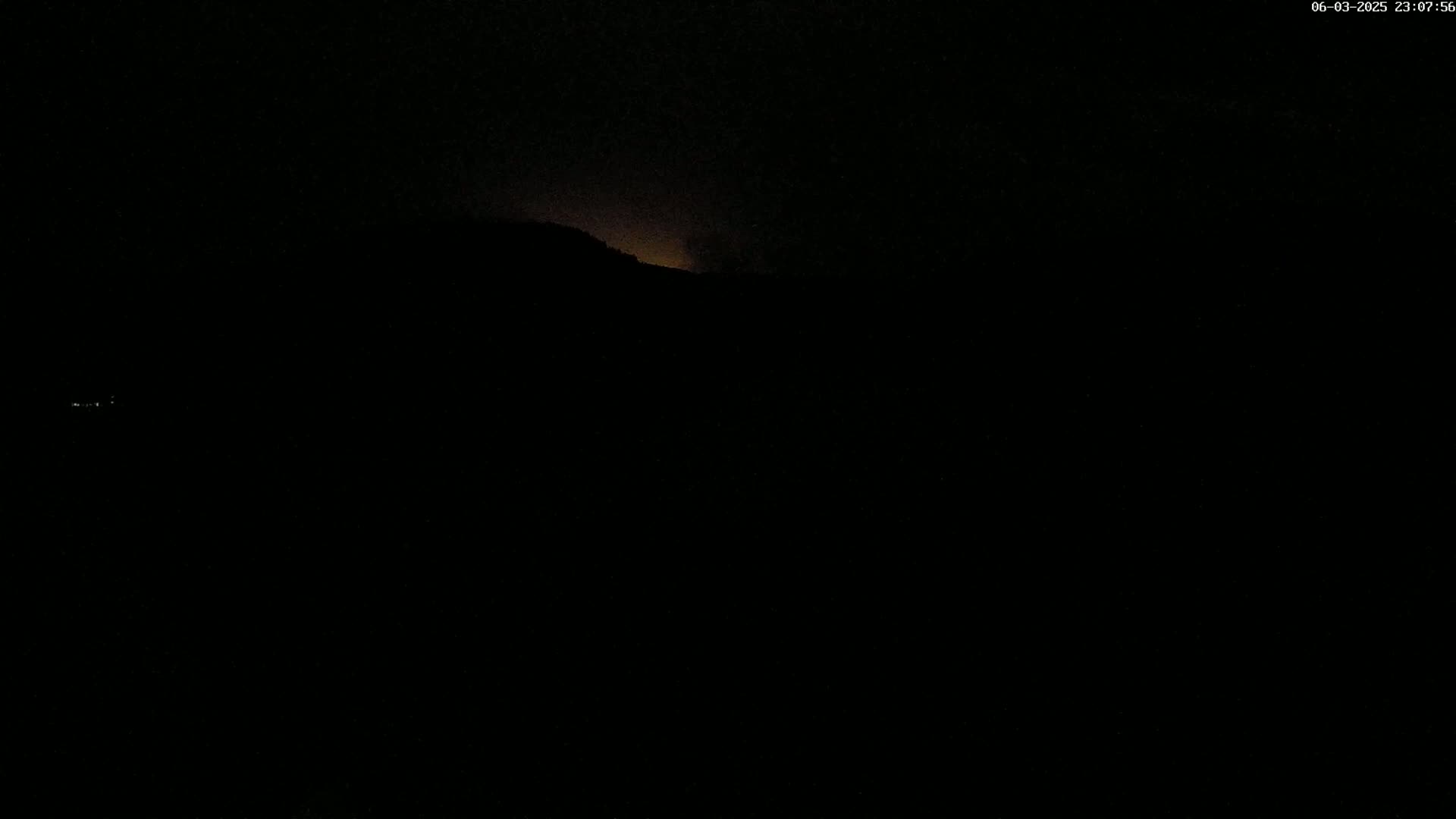 A dark, nighttime scene shows a dimly lit mountain range under a mostly clear sky.