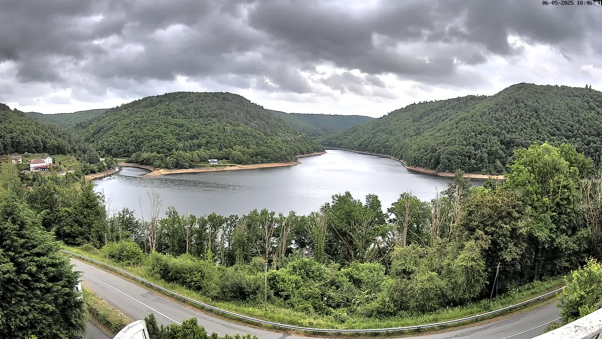 A winding lake nestled between lush green hills under a cloudy sky is viewed from a high vantage point above a road.