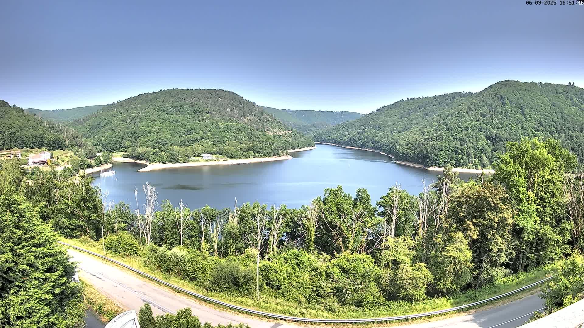 A winding road overlooks a calm, blue lake nestled within a valley of lush green hills under a clear blue sky.