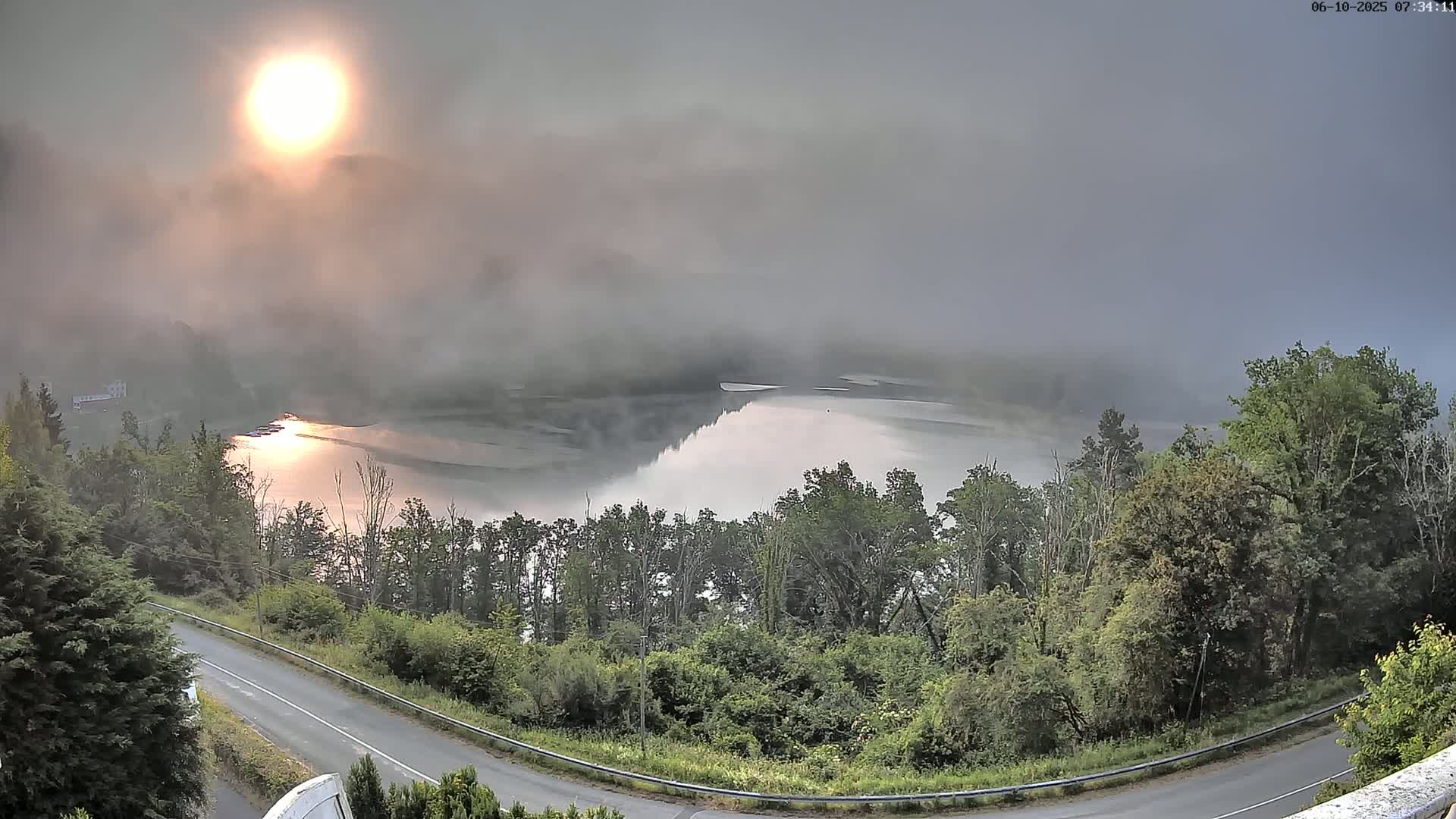 A foggy sunrise illuminates a calm lake nestled in a green, wooded valley, viewed from above a winding road.