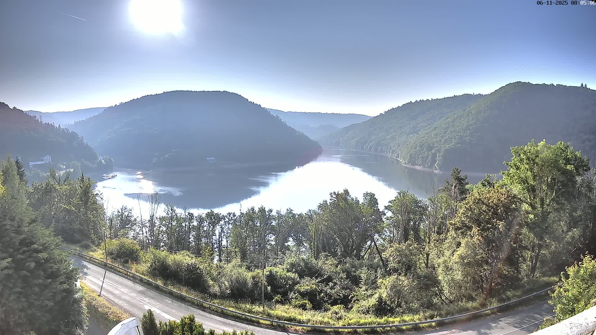 A bright, sunny day reveals a calm lake nestled between lush green hills, with a road curving gently through the foreground.