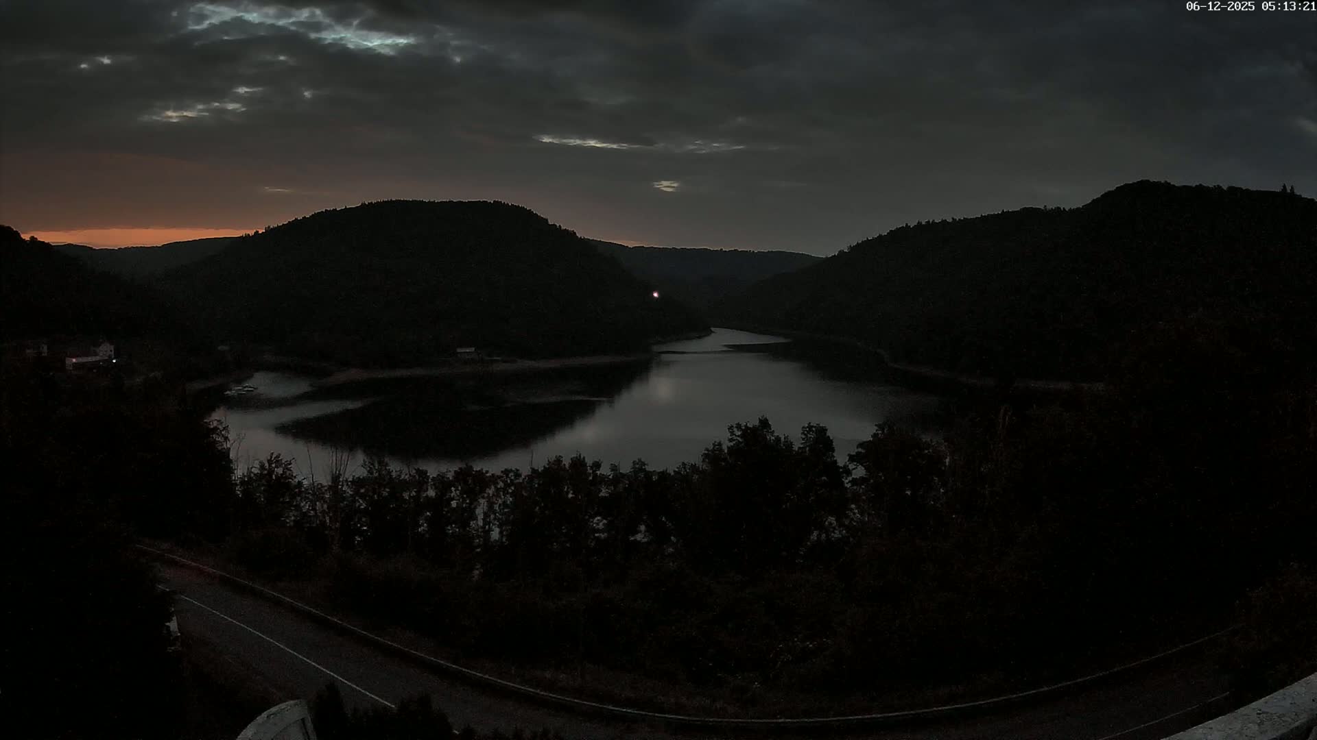 A dark, calm lake nestled between silhouetted hills under a cloudy, pre-dawn sky.