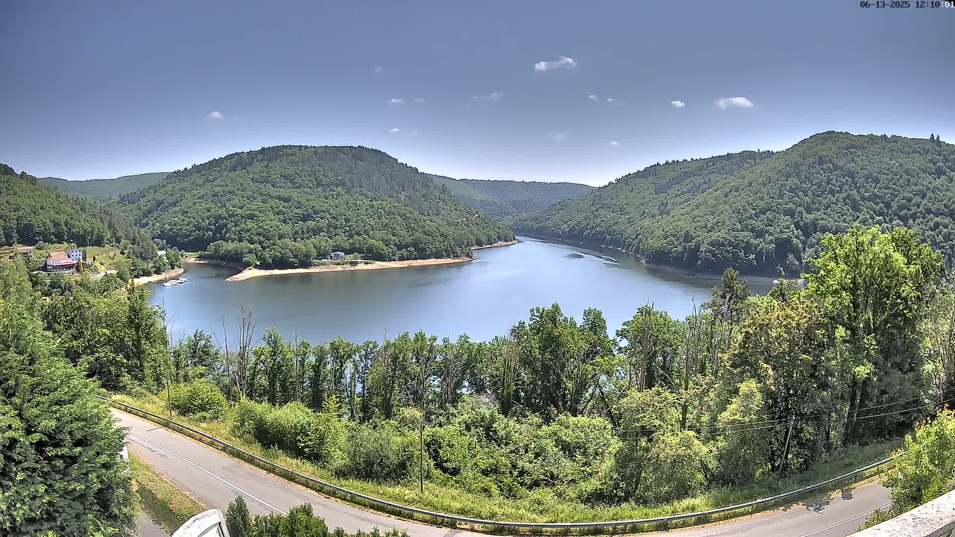 A winding road overlooks a calm lake nestled between lush green hills under a mostly clear blue sky.