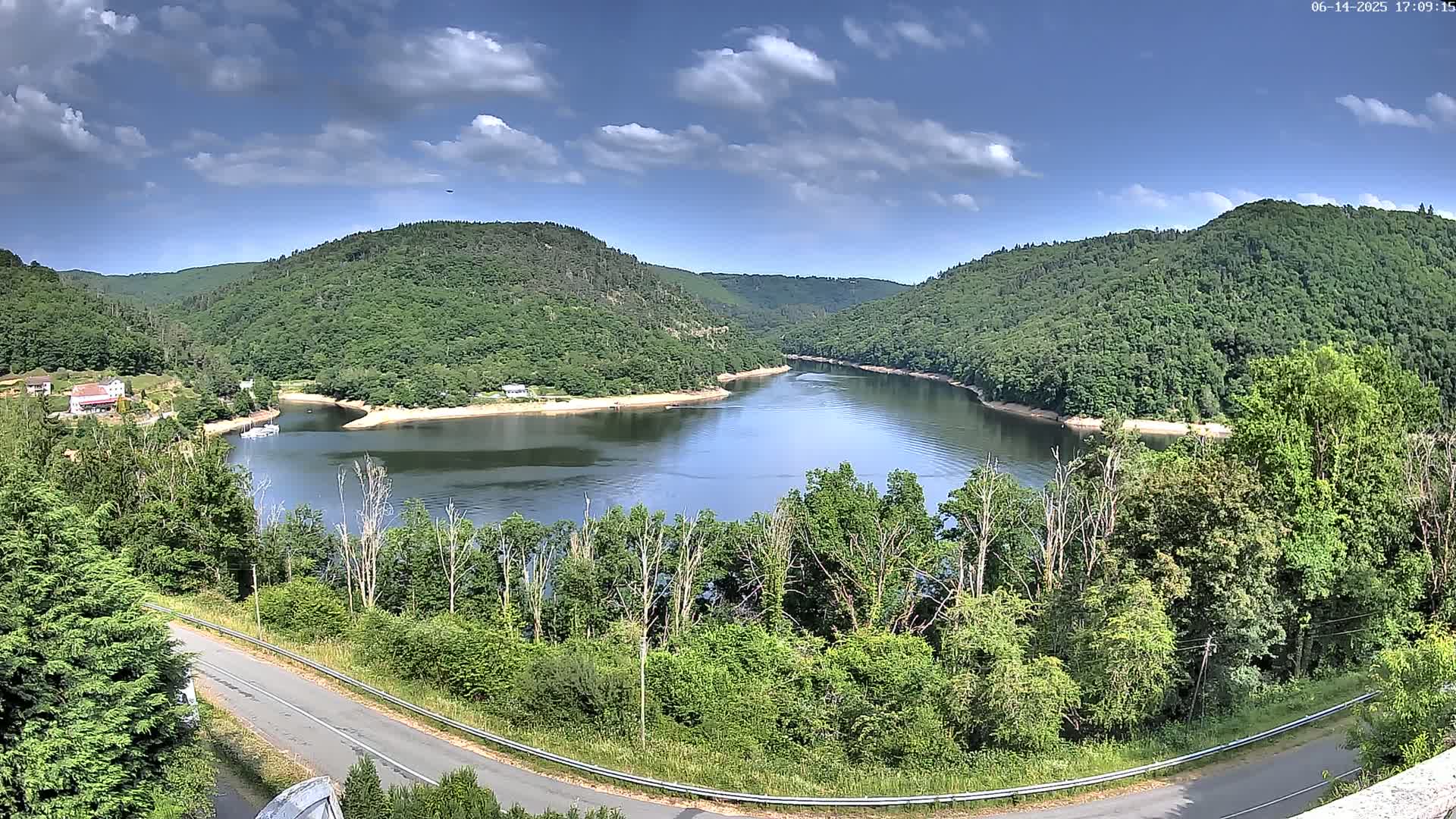 A partly cloudy sky overlooks a calm lake nestled between lush green hills, with a road curving alongside the water's edge.