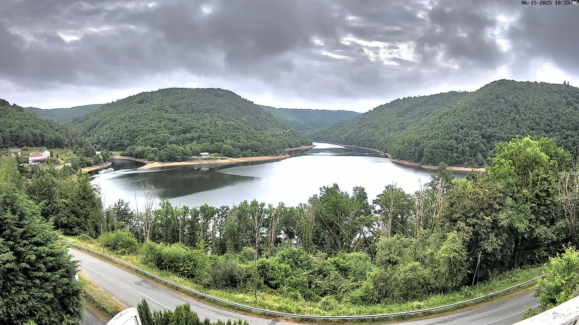 A winding road overlooks a calm lake nestled in a valley surrounded by lush green hills under a cloudy sky.