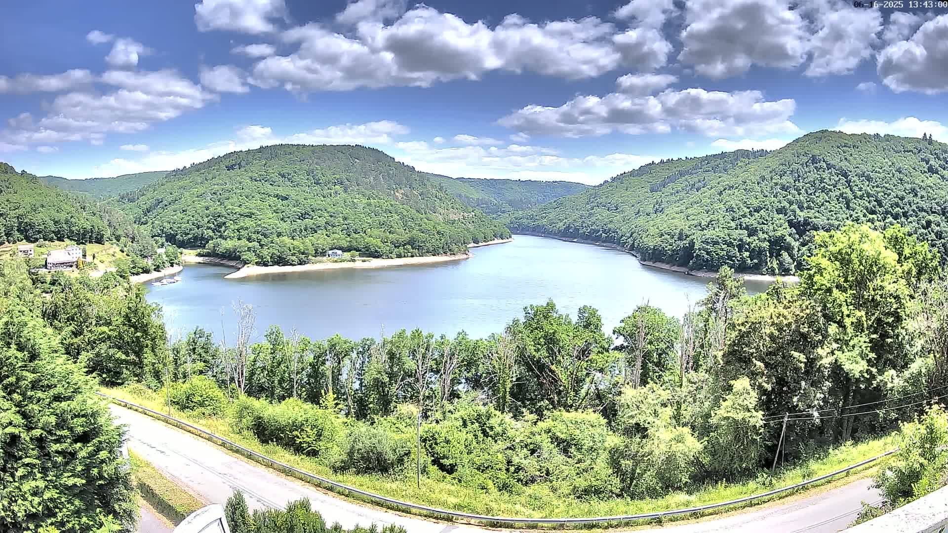 A partly cloudy sky overlooks a calm, curved lake nestled in a valley of lush green hills and trees.