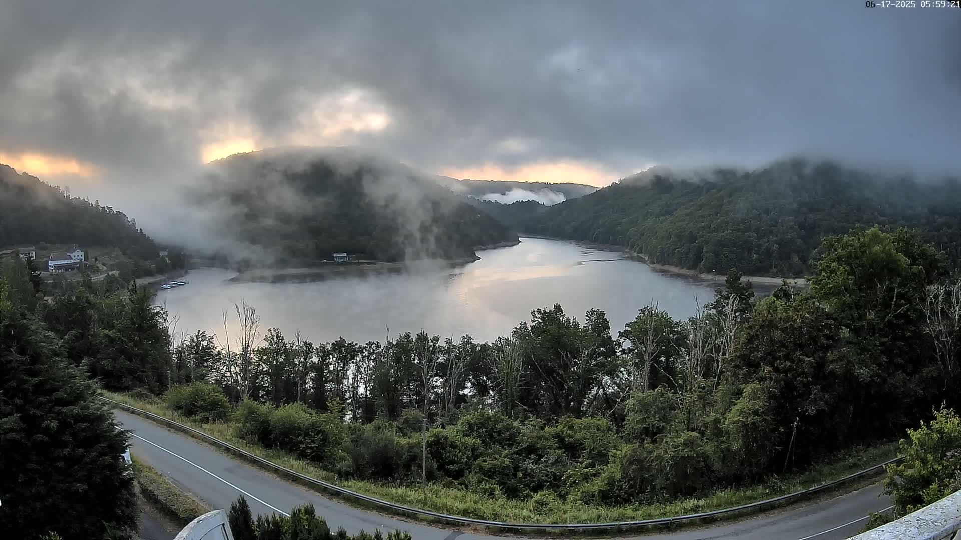 A winding road overlooks a lake nestled in a valley shrouded in morning fog, with the sun partially visible through the clouds.