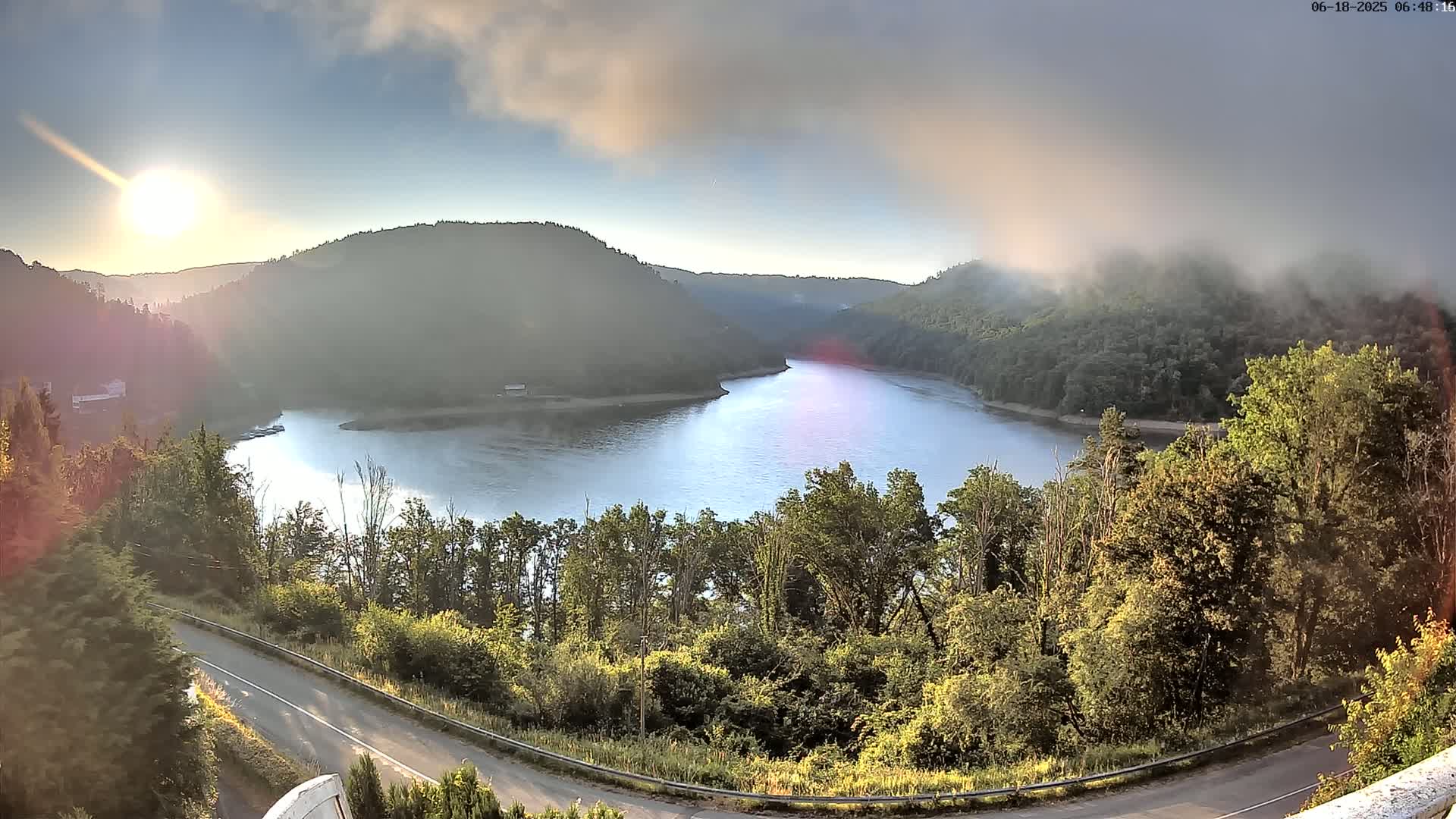 A calm lake nestled in a valley between sunlit, fog-covered hills, viewed from a winding road above.