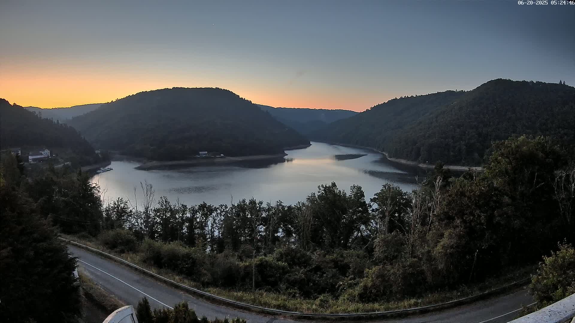 A calm lake nestled between dark green hills at sunrise, with a paved road curving along the lower right.