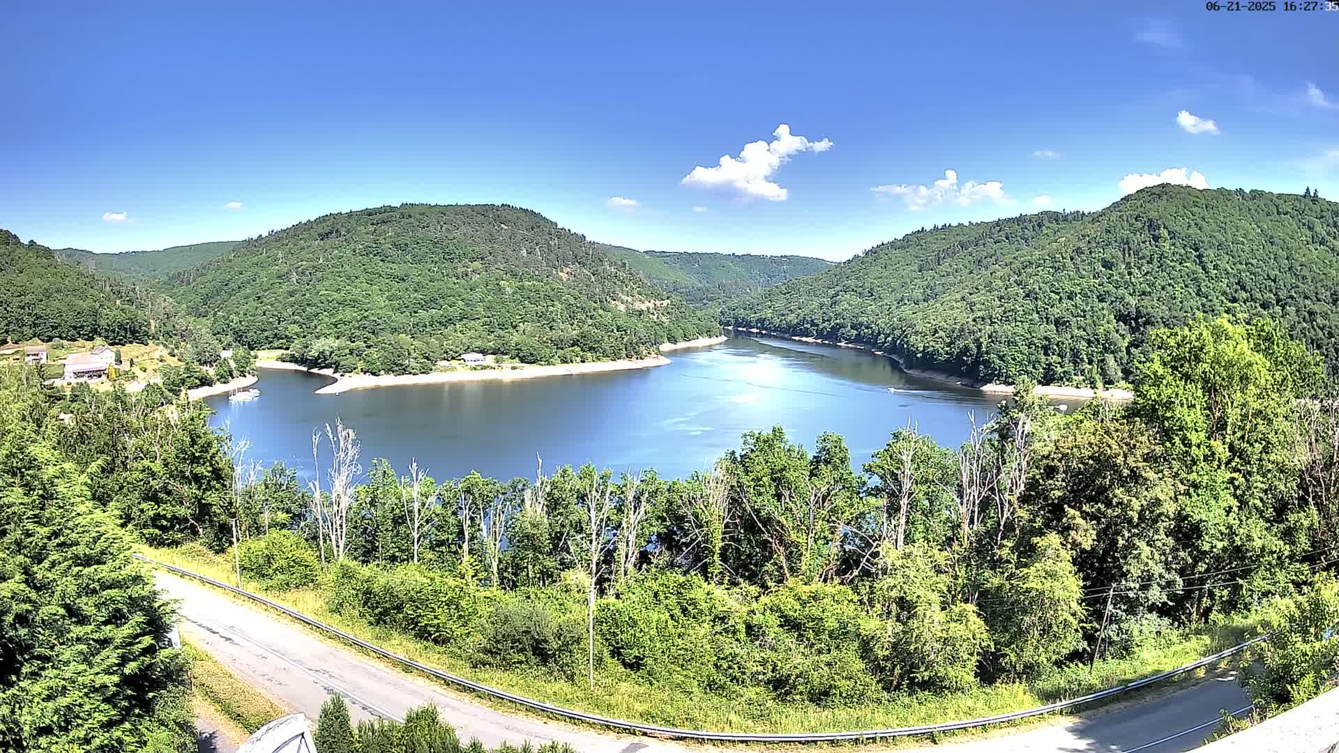 A winding road overlooks a calm lake nestled in a valley of lush green hills under a mostly sunny sky.