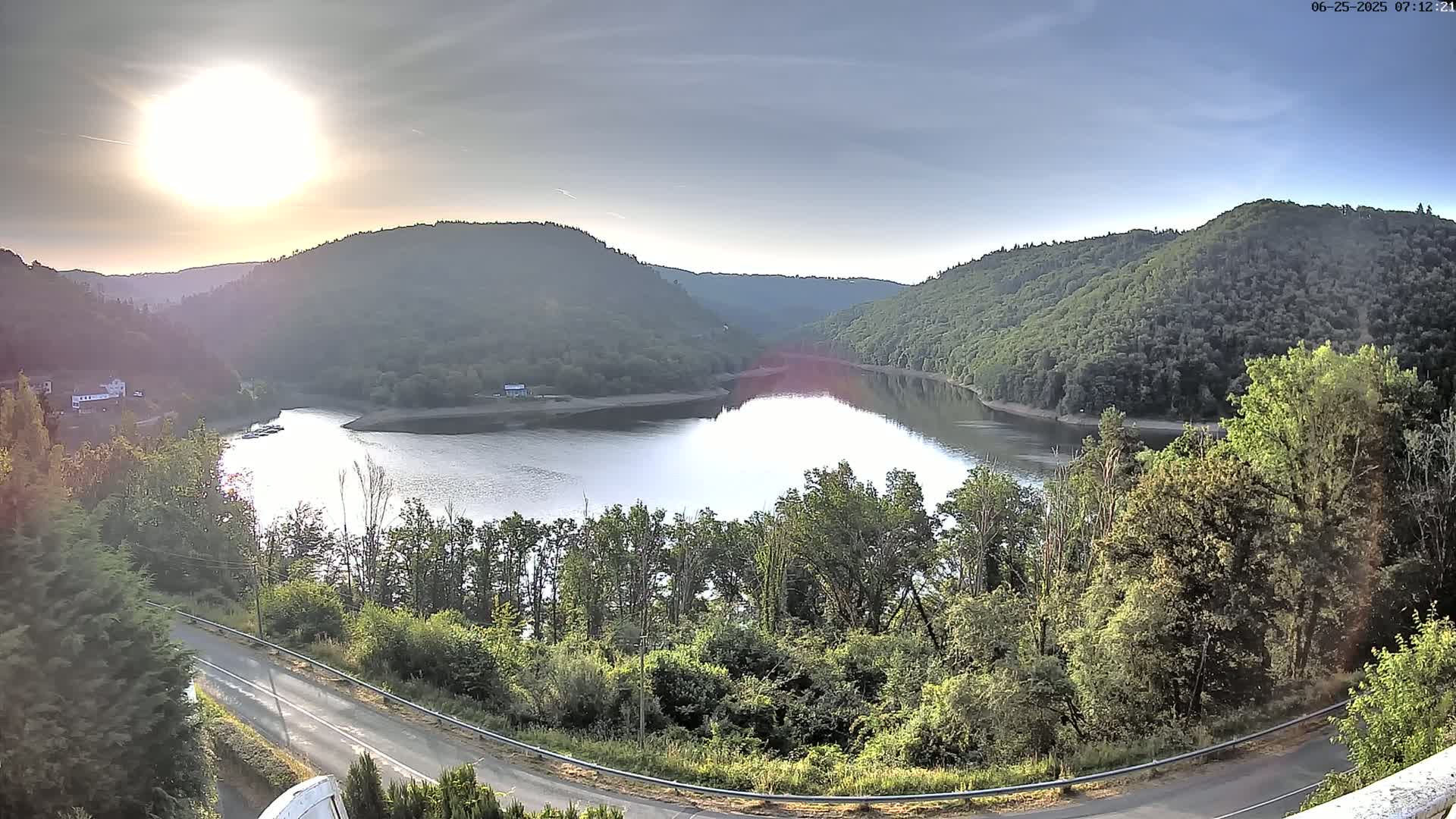 A calm lake nestled between verdant hills is viewed from an elevated position on a sunny morning.