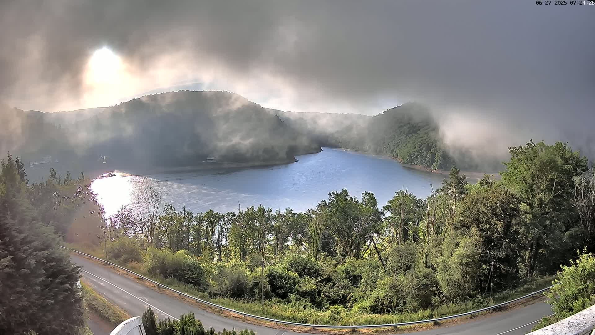 A calm lake nestled in a valley shrouded in morning mist, overlooked by a winding road bordered by lush green trees.