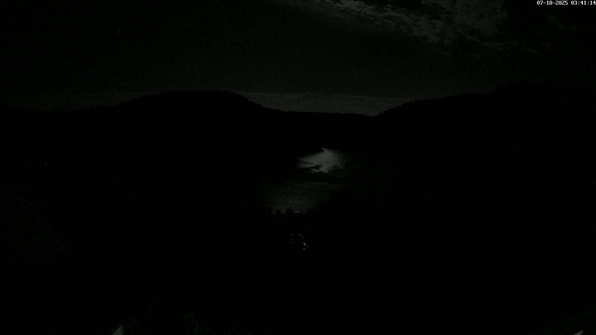 A nighttime view shows a dark, moonlit body of water nestled between dark hills under a cloudy sky.