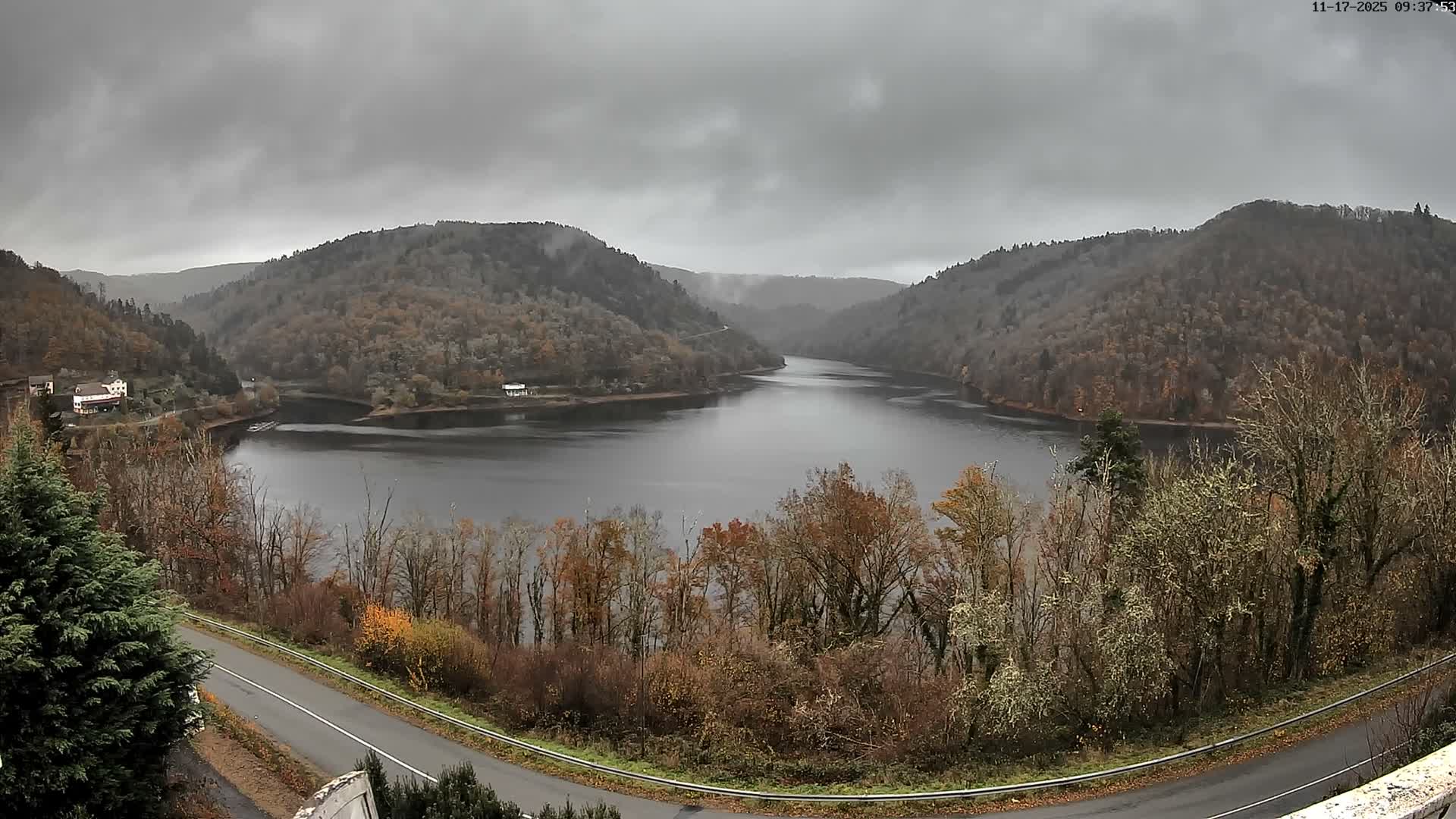 A dark body of water winds through a valley bordered by tree-covered hills displaying autumn foliage, with a road in the foreground and a few scattered buildings, all beneath a heavily overcast gray sky.