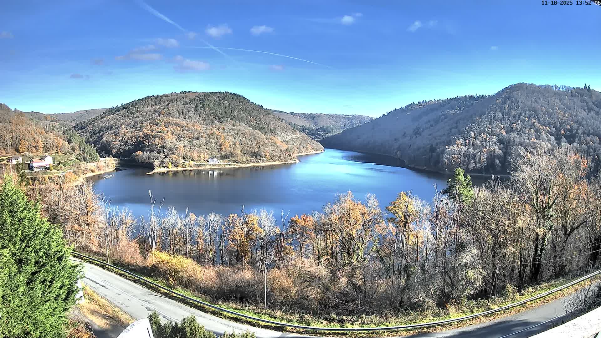 A dark body of water winds through a valley bordered by tree-covered hills displaying autumn foliage, with a road in the foreground and a few scattered buildings, all beneath a heavily overcast gray sky.